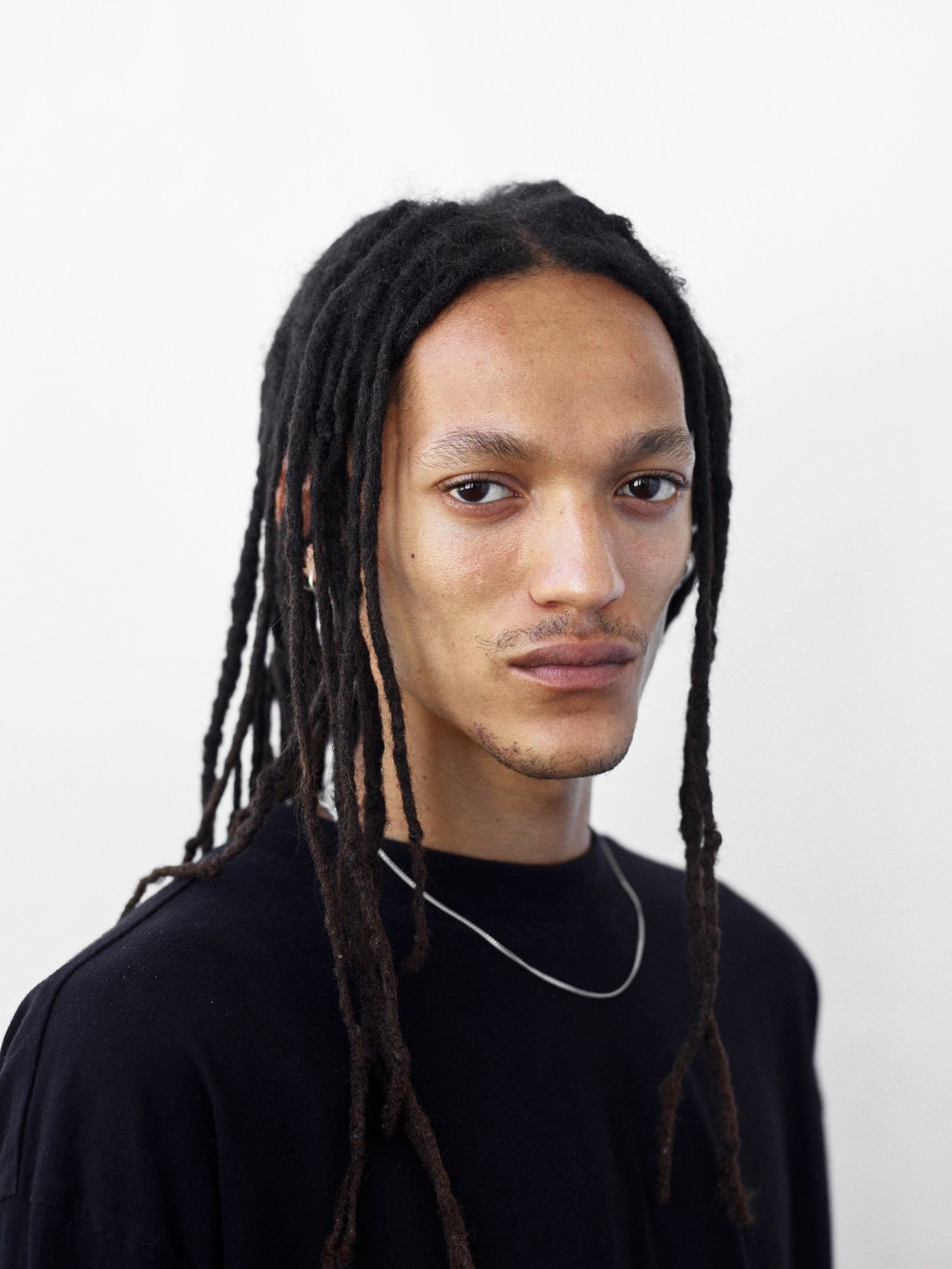 A man with long black dreads wearing a black top and silver chain necklace