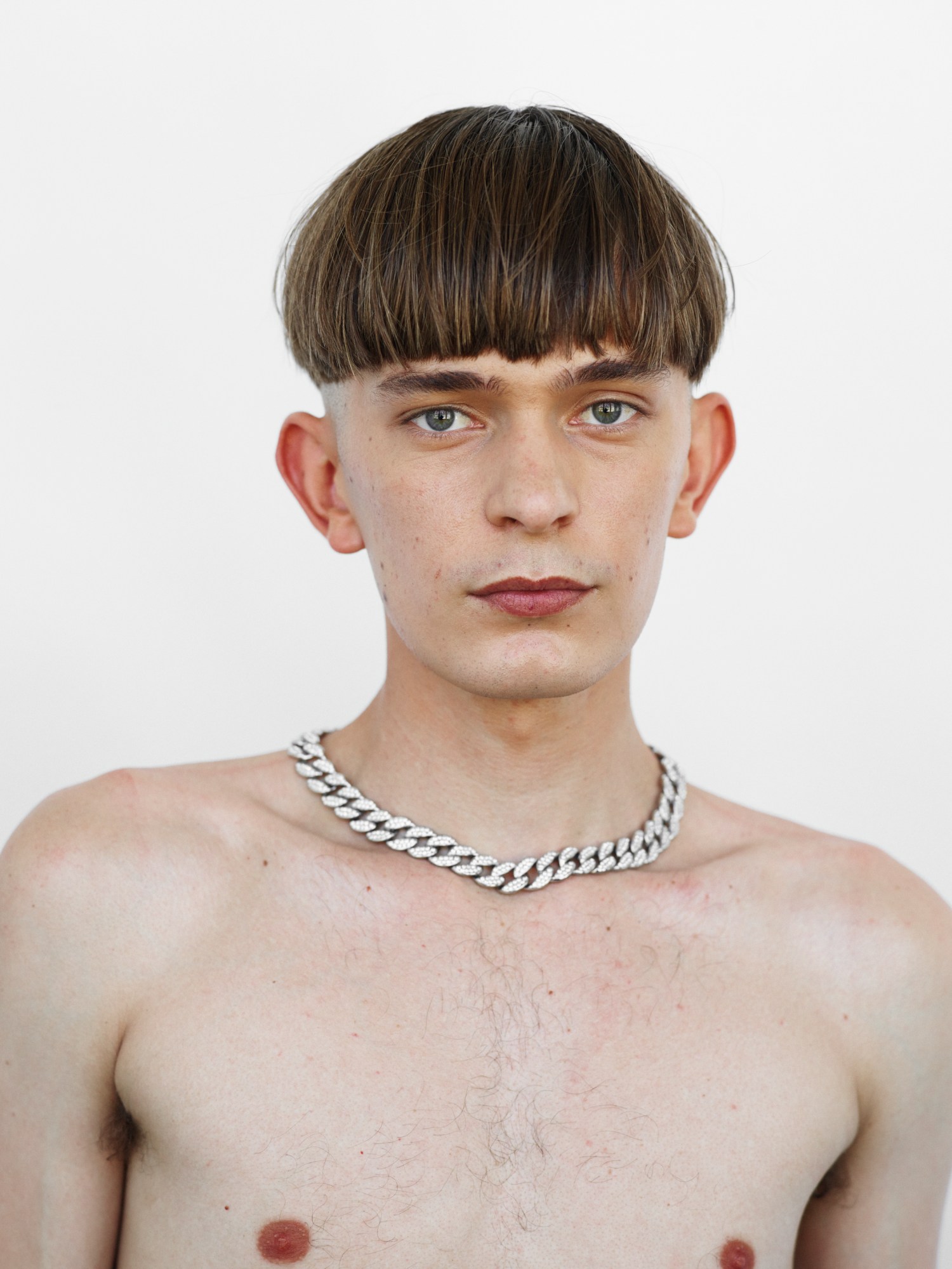 A topless man with brown block fringed bowl cut wearing a silver chunky chain necklace