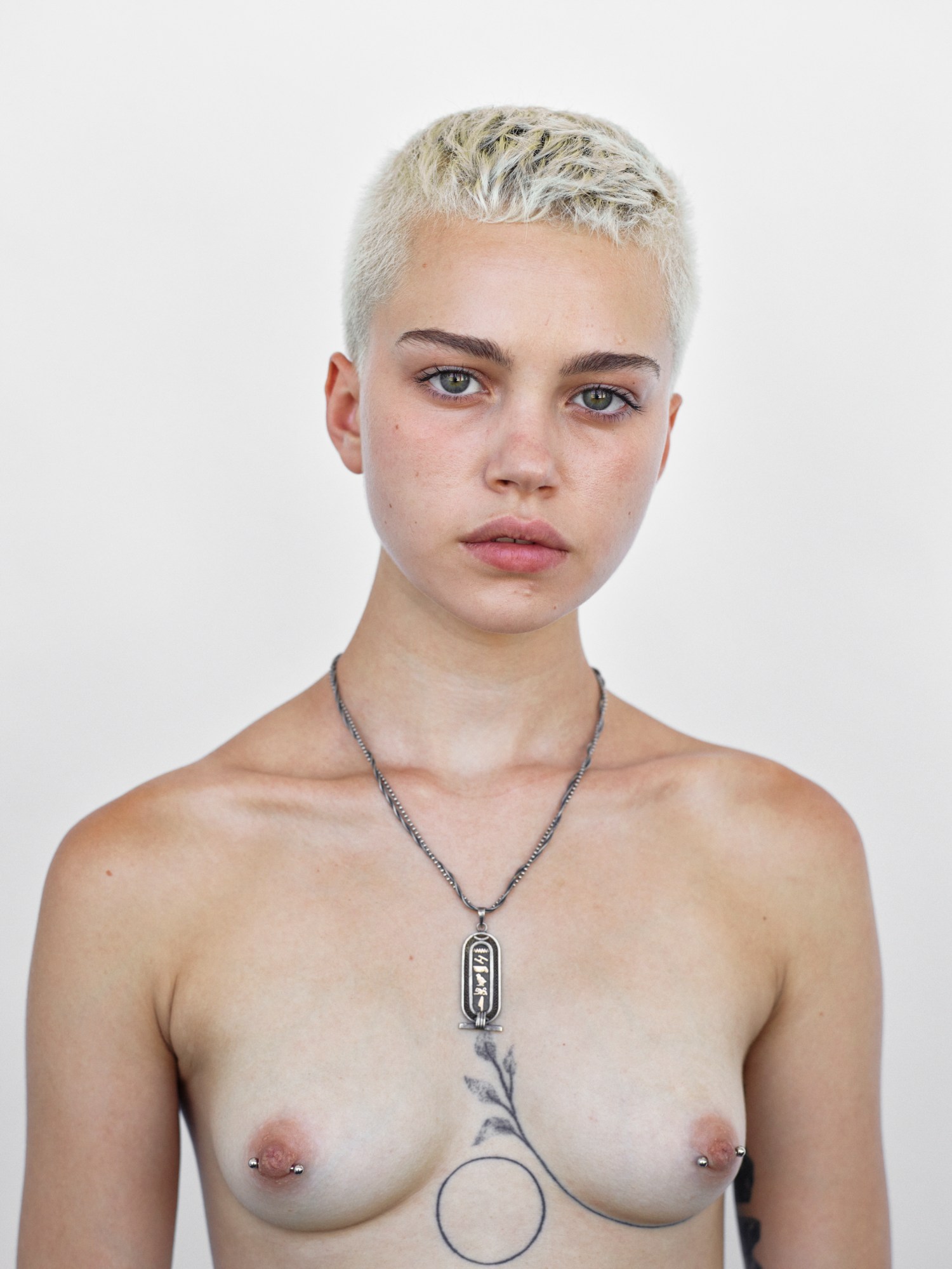 A topless woman with short white blonde hair and underboob tattoos wearing nipple piercings and a silver chain.