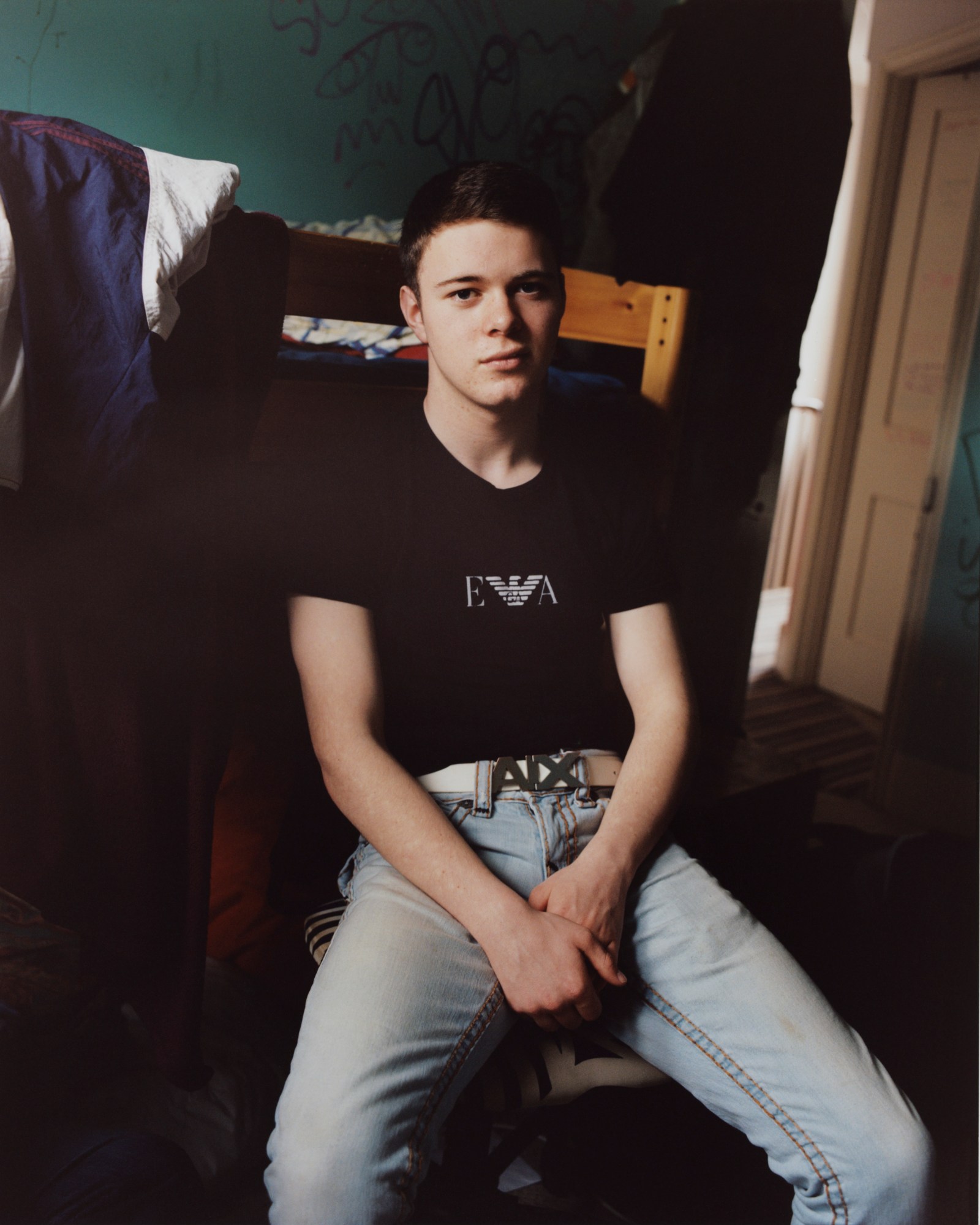 boy in light blue jeans and emporio Armani t-shirt sits on a chair in his room