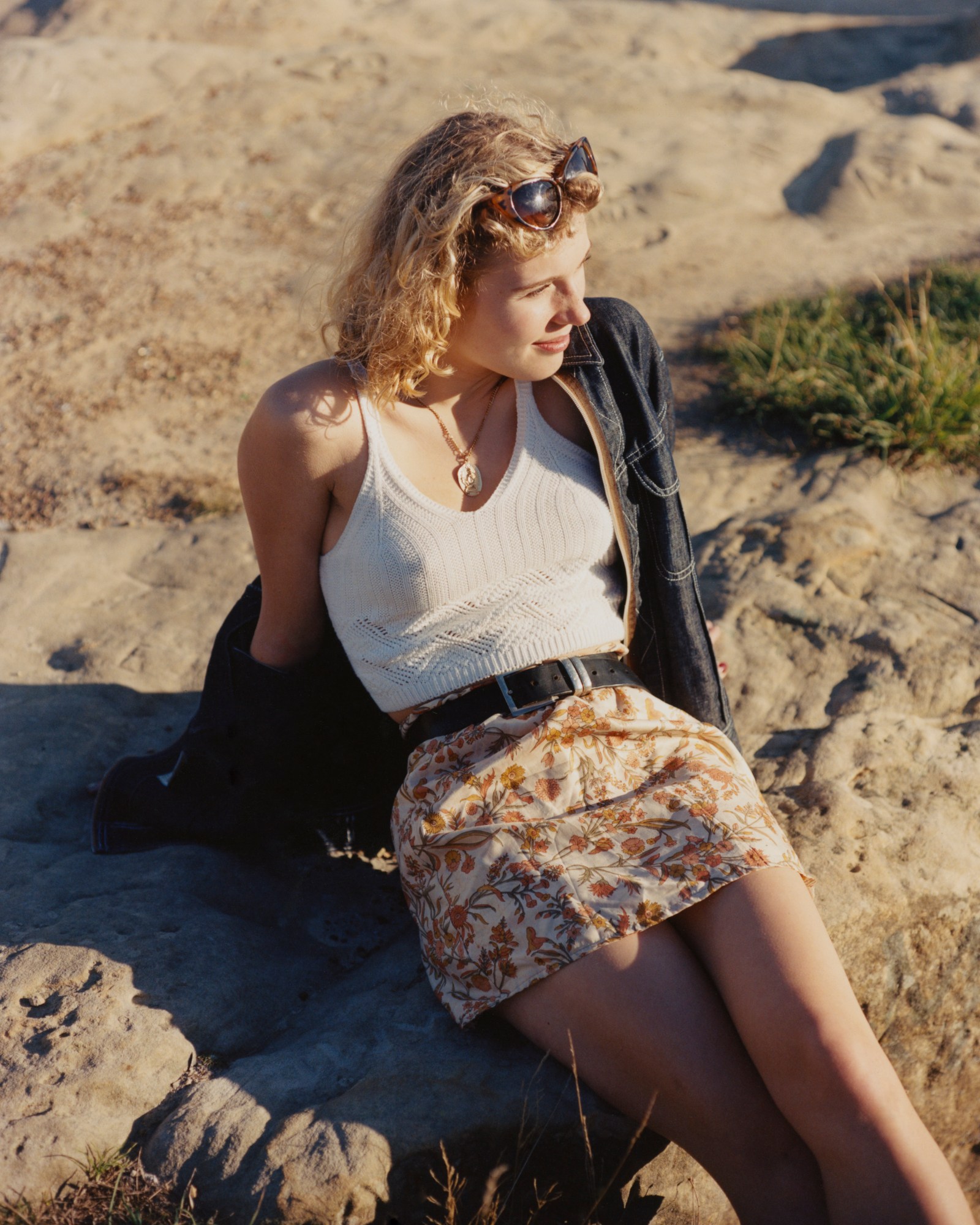 Girl in floral skirt, white knitted vest and denim jacket sits on the rocks.