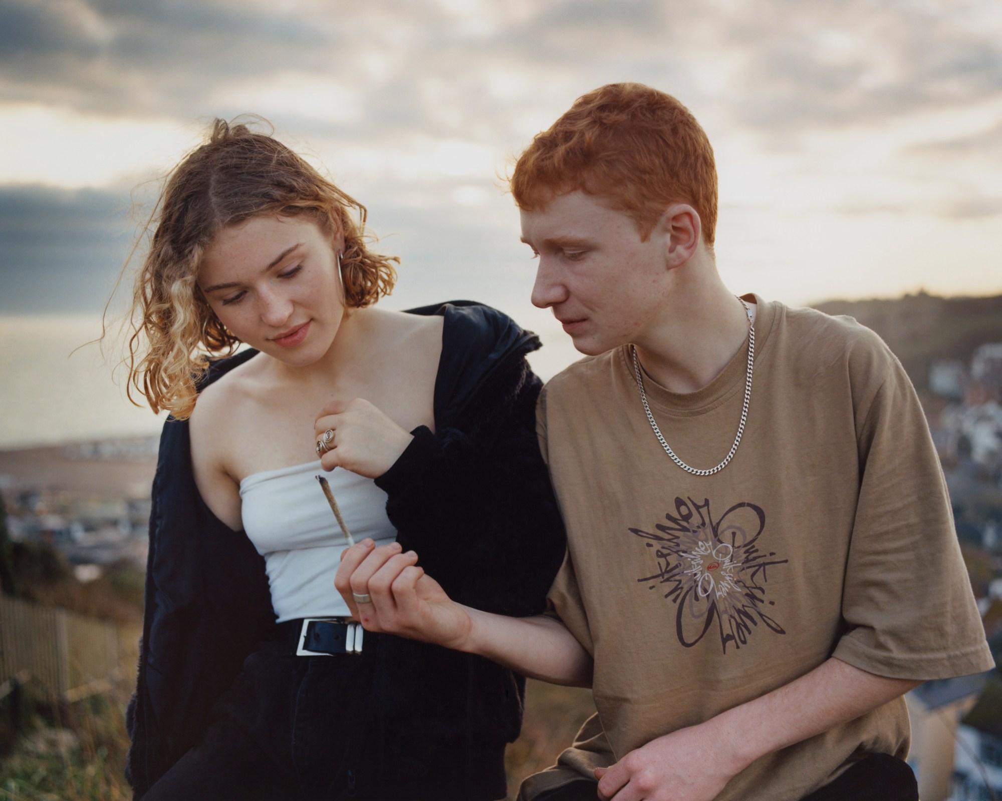 A boy passes a girl his cigarette while sitting on a cliff top.