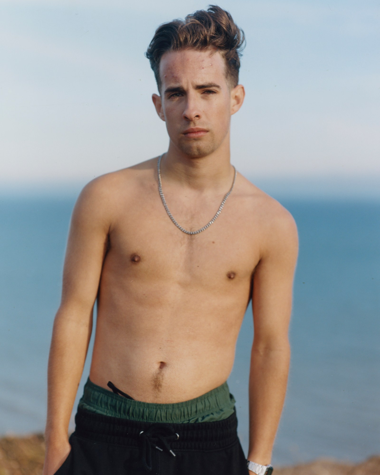 Shirtless boy standing by the ocean wearing swim shorts and joggers on top and a silver chain.