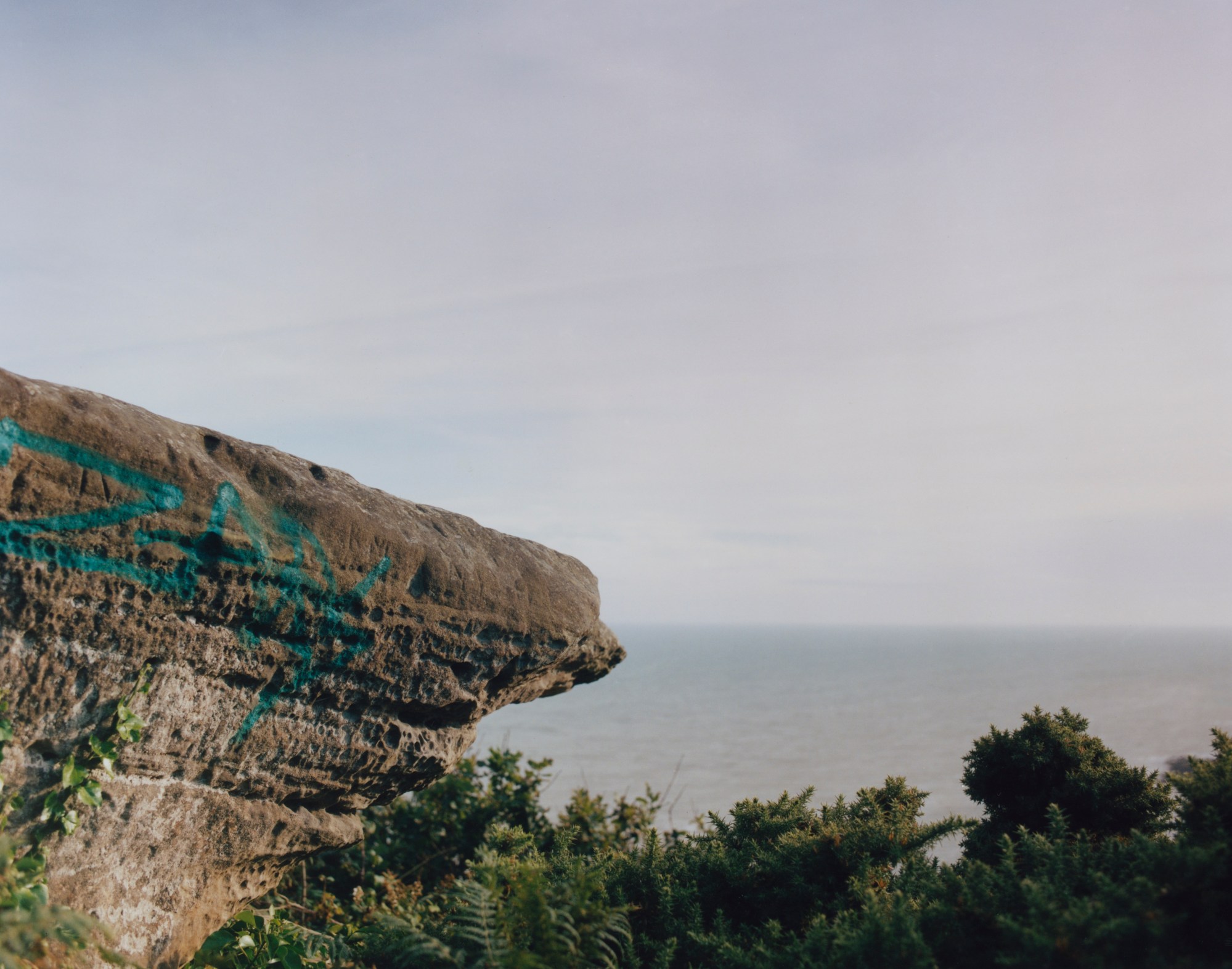 Edge of a cliff with blue graffiti on it. Ocean and bushes are in the background.