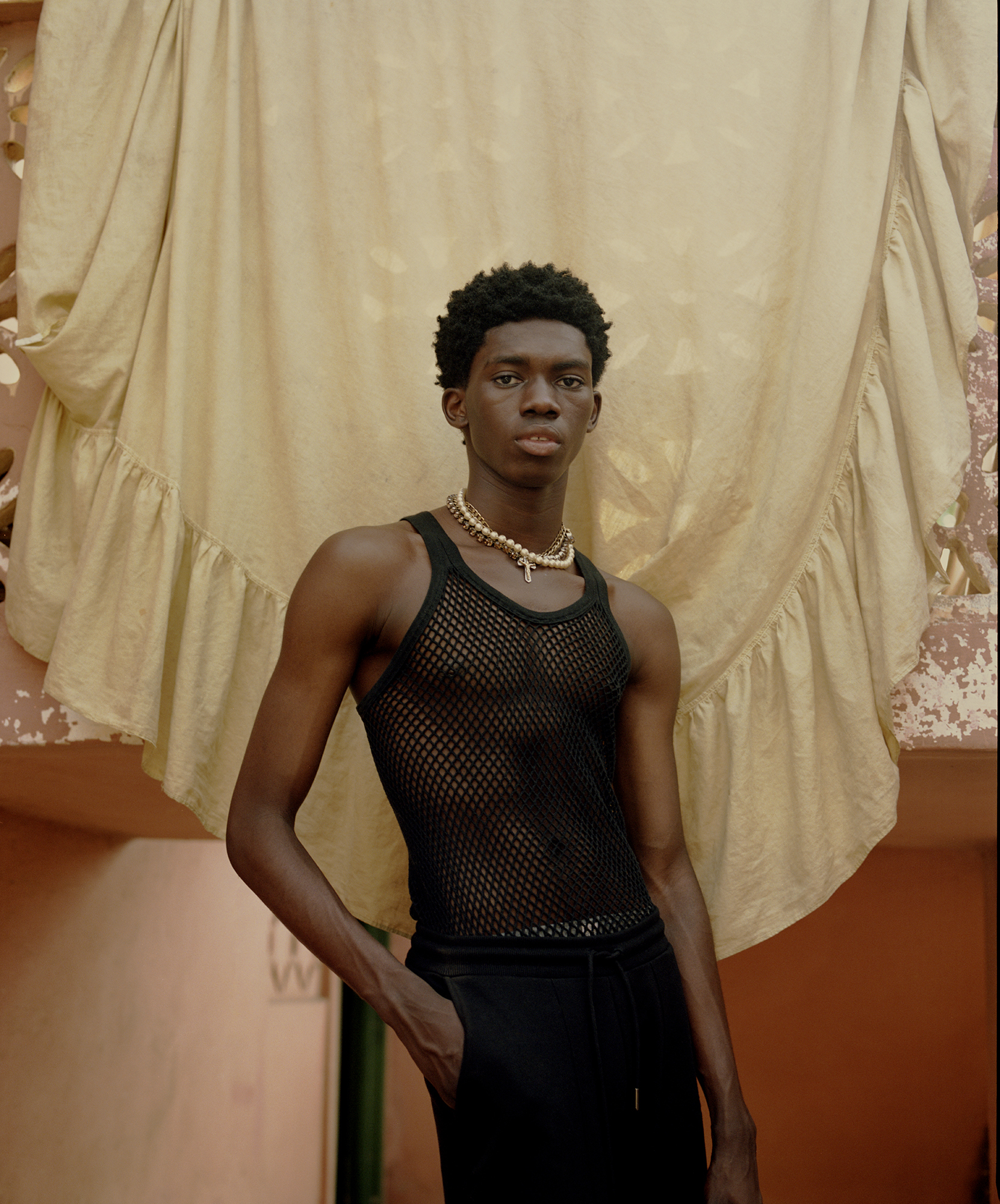 a boy posing in a net tank top and pearls in front of a tapestry