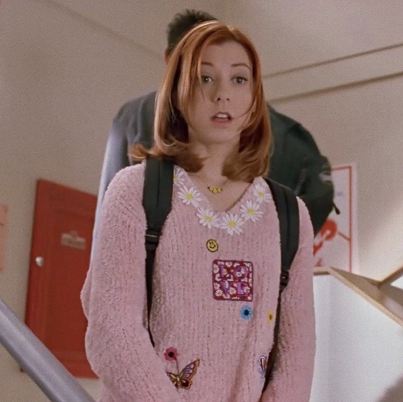 Willow walks down the school stairwell in a pink jumper with a waist chain print along the collar and butterfly, flower, smiley and LOVE patches across the front. She also has a black backpack.