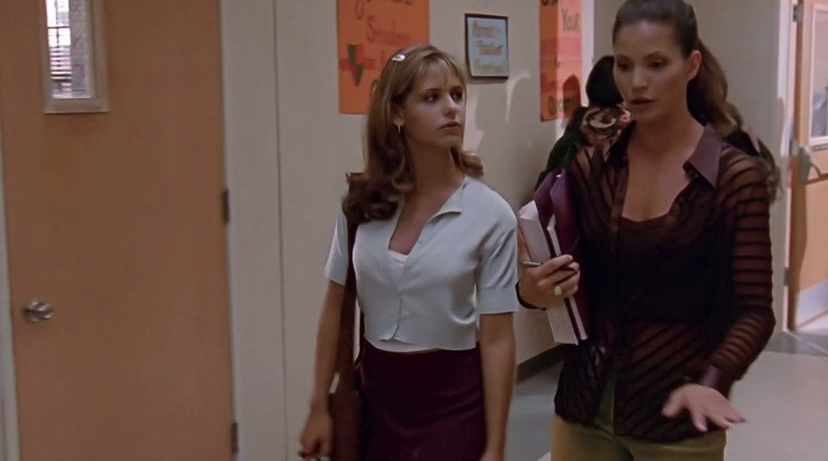 Buffy and Cordelia walk through the school hallway. Buffy wears a burgundy mini skirt, white vest top, blue cardigan and a barrette while Cordelia wears lime trousers, a burgundy vest top and a sheer burgundy striped shirt. She also carries her school books.