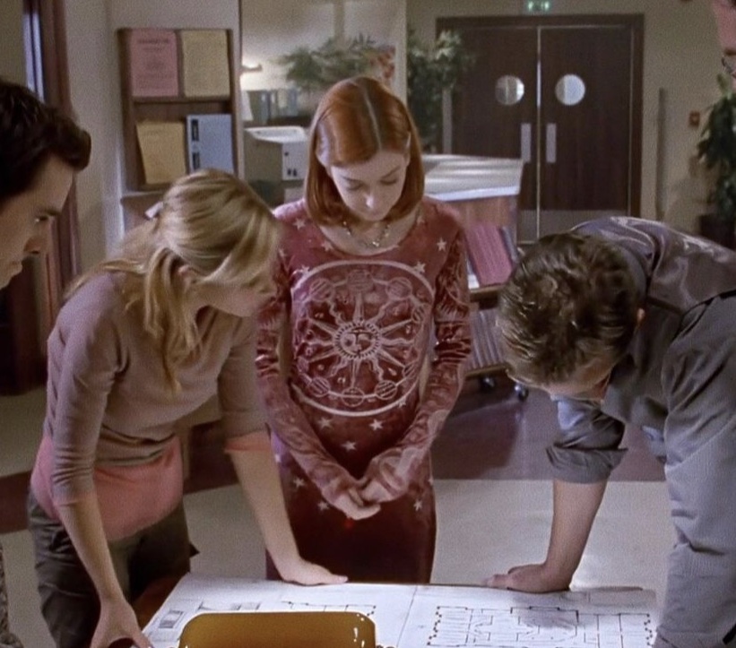 Buffy, Willow, Xander and Giles look at a map blueprint in the school library. Willow wears a red velvet dress with an astrology print across the front and sleeves.