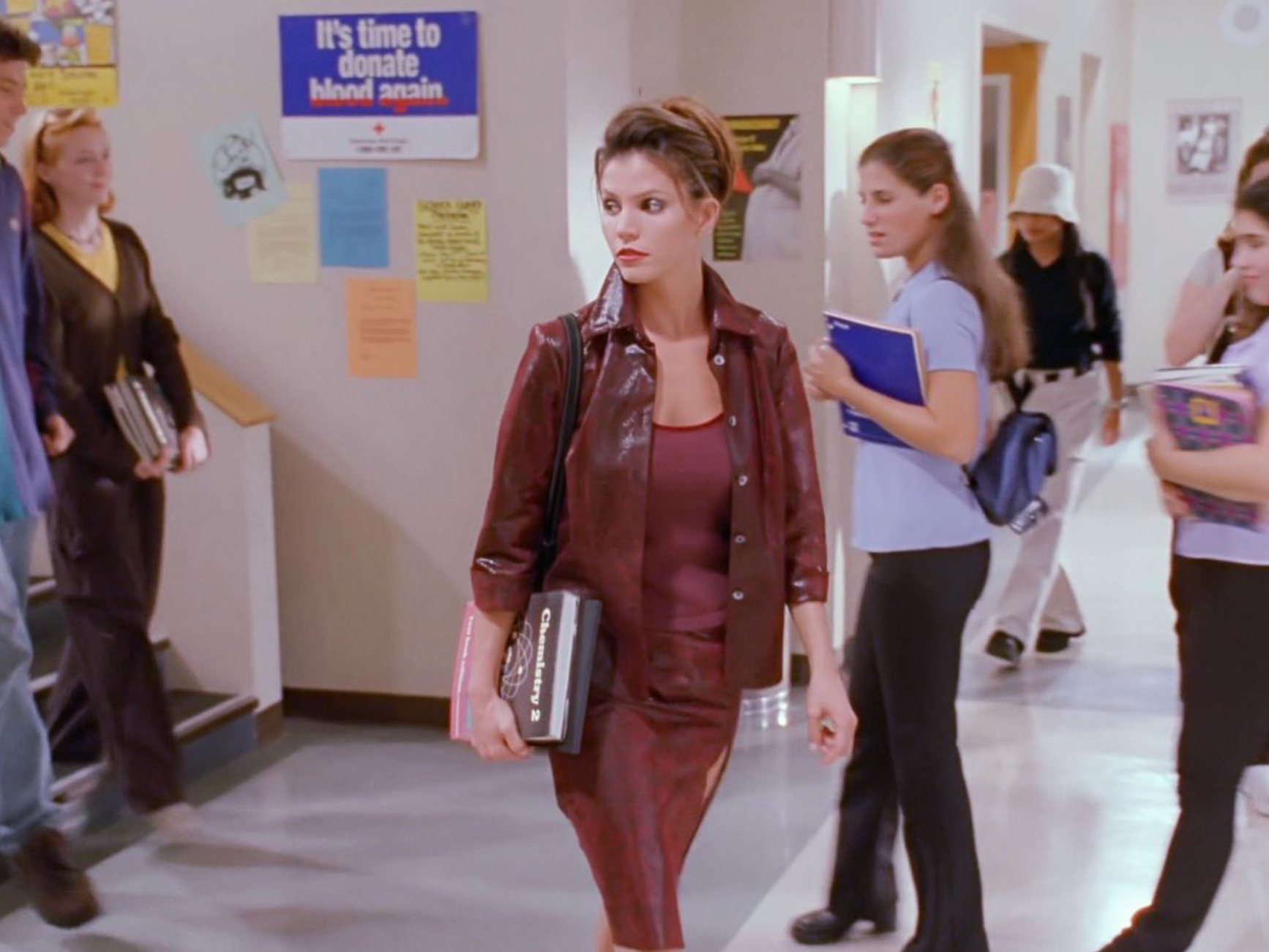 Cordelia in the school halls wearing a mahogany dress, mahogany jacket and carrying books.