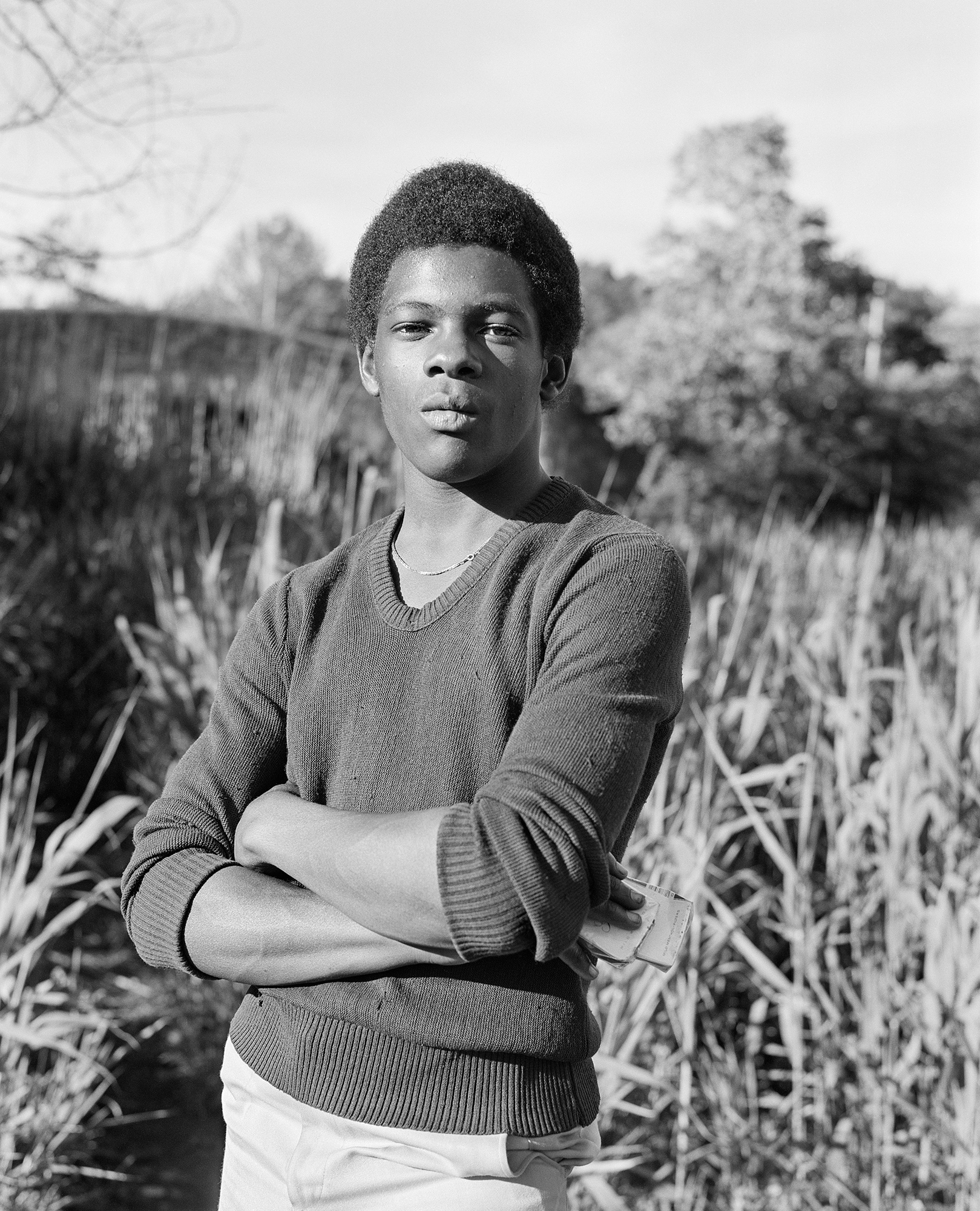 a young man stands with his arms folded against long grass