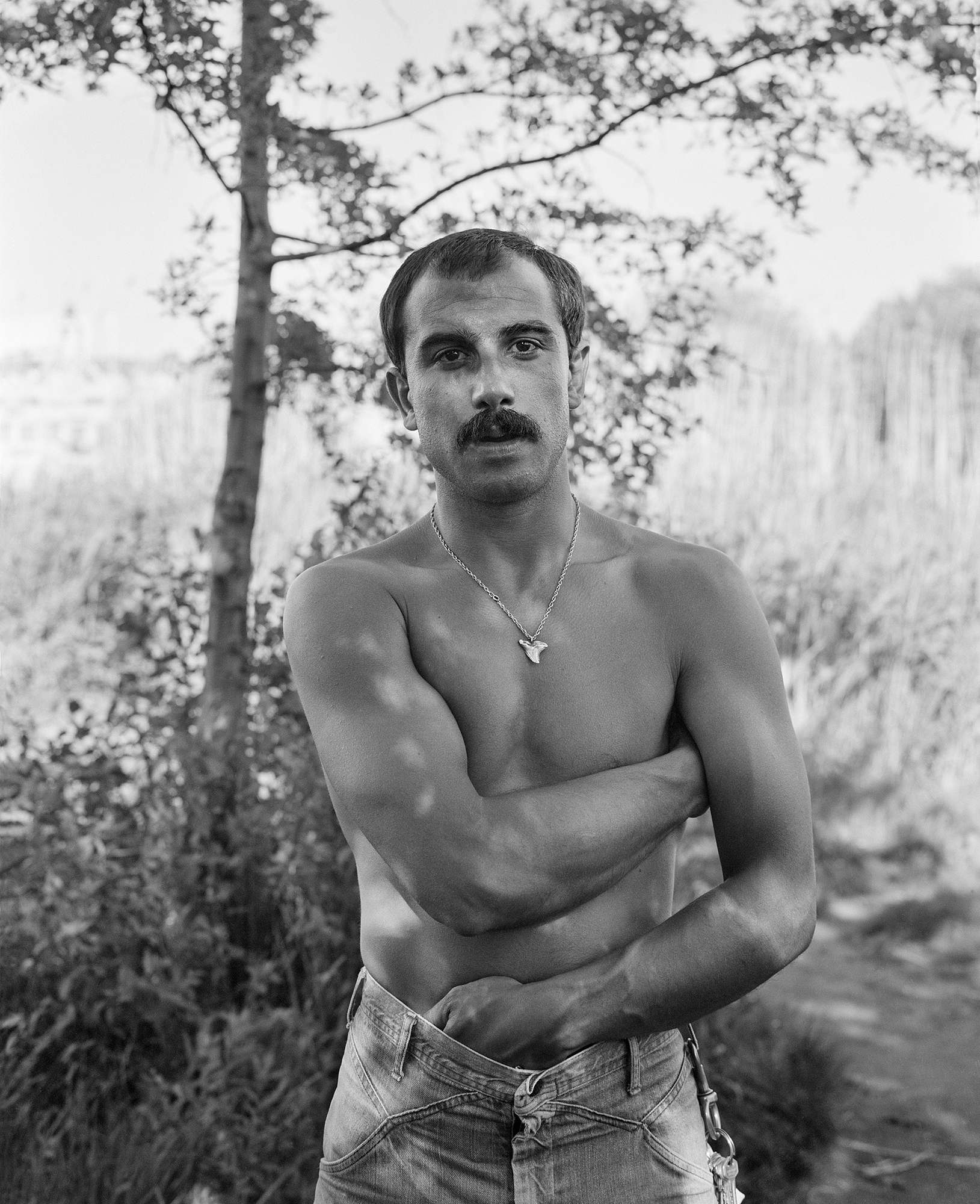 a topless man stands in the shade surrounded by trees and long grass