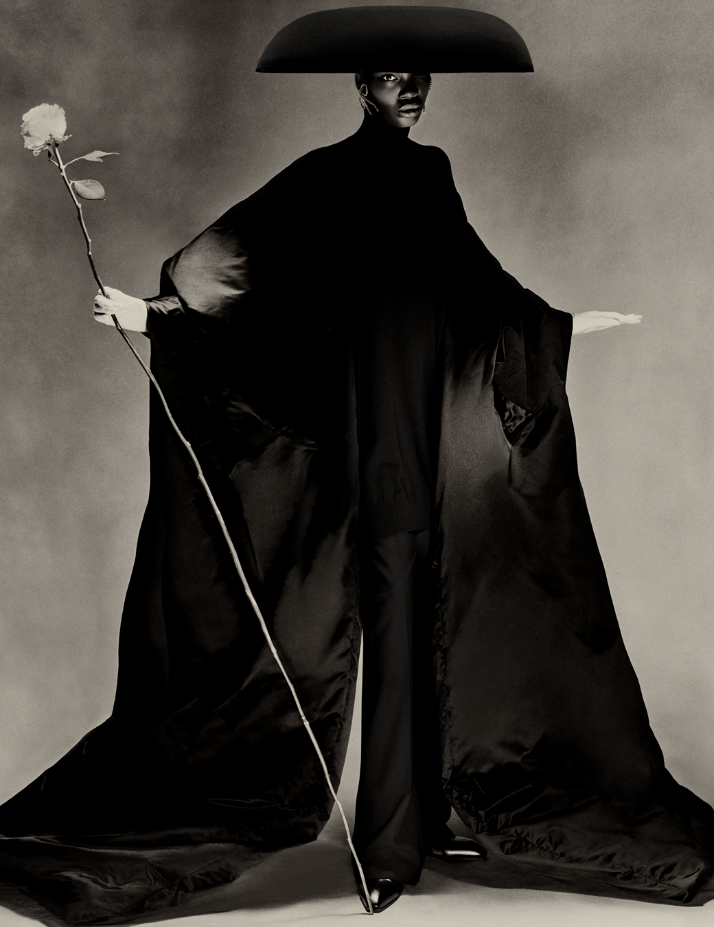 model wears a black dress with draping sleeves and black trousers with an umbrella hat all by balenciaga and a giant stem rose in her hand