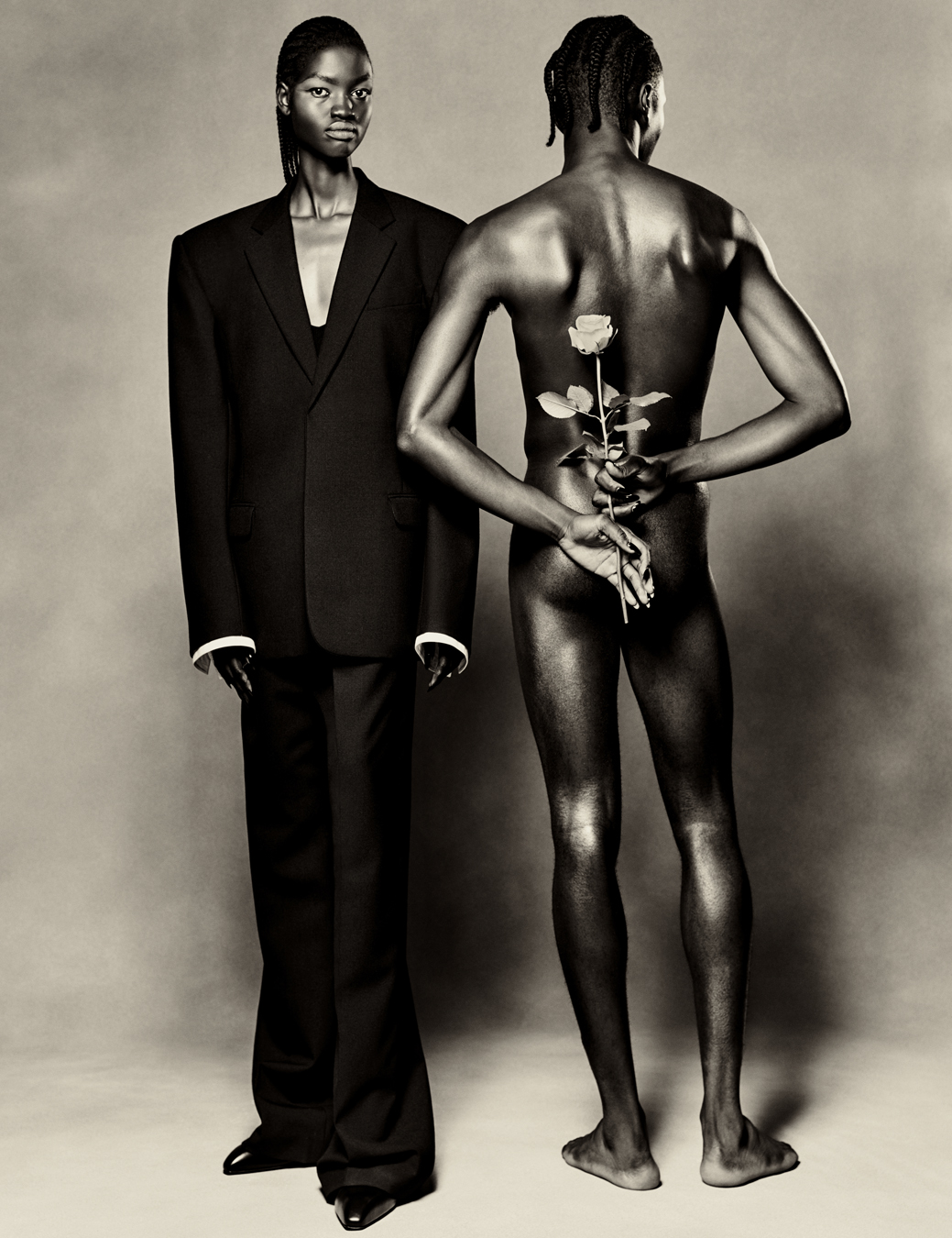 One model wears two piece suit by balenciaga. The other is naked facing the wall and holds a rose.