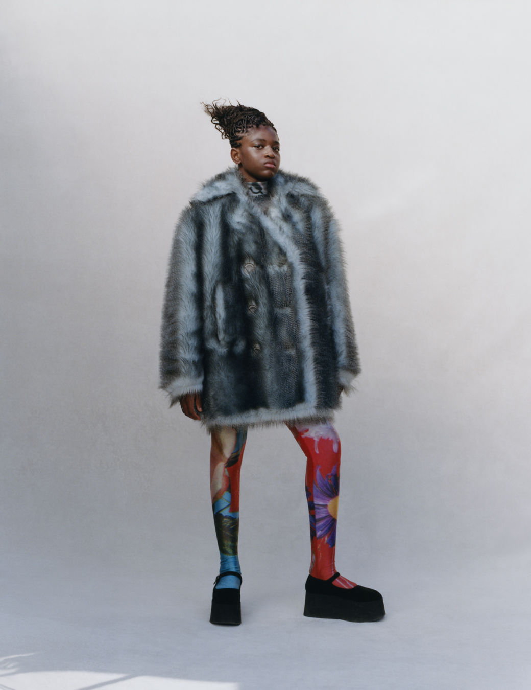 Zaya stands in a grey room wearing a fluffy coat, colourful floral tights and black platform buckles.