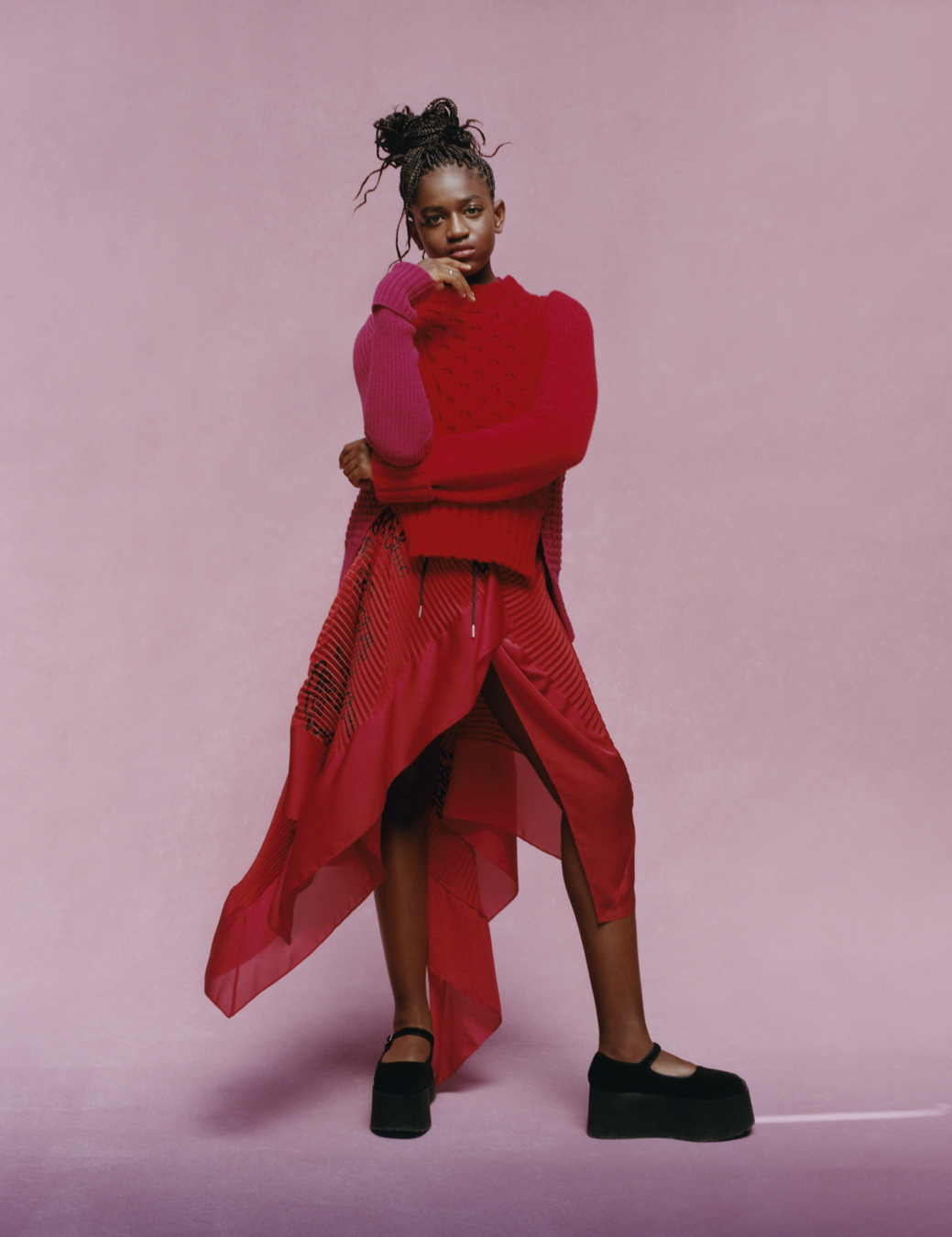 Zaya wears a red skirt and red hoodie with black platform buckle shoes standing in a pink room.