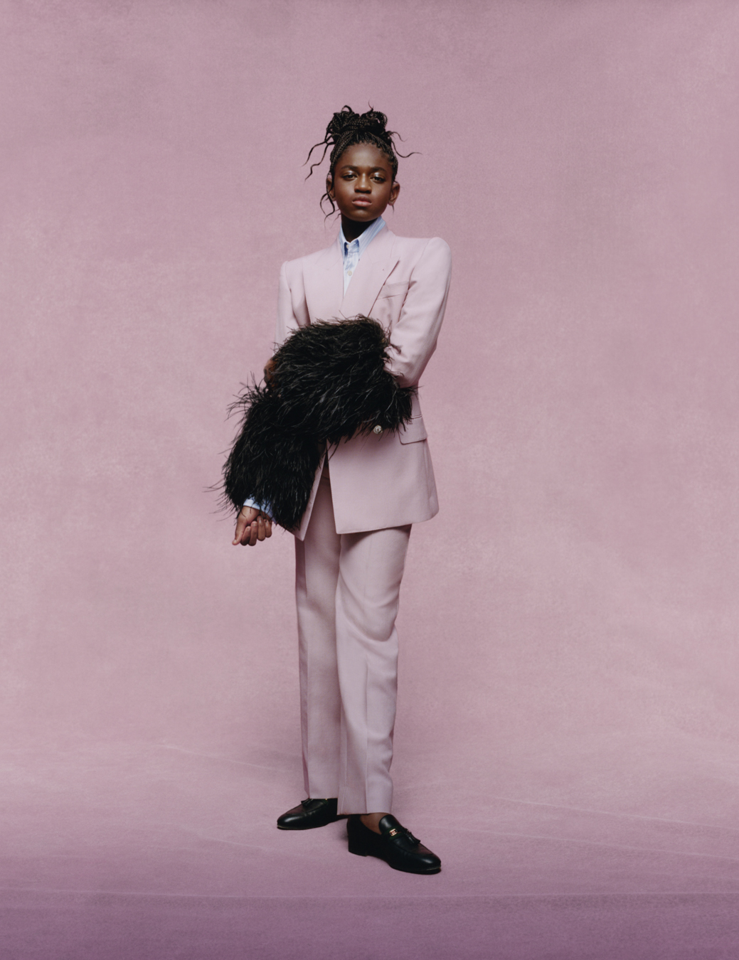 Zaya Wade wearing a pink suit, black feathery sleeves and black shoes standing in a pink room.