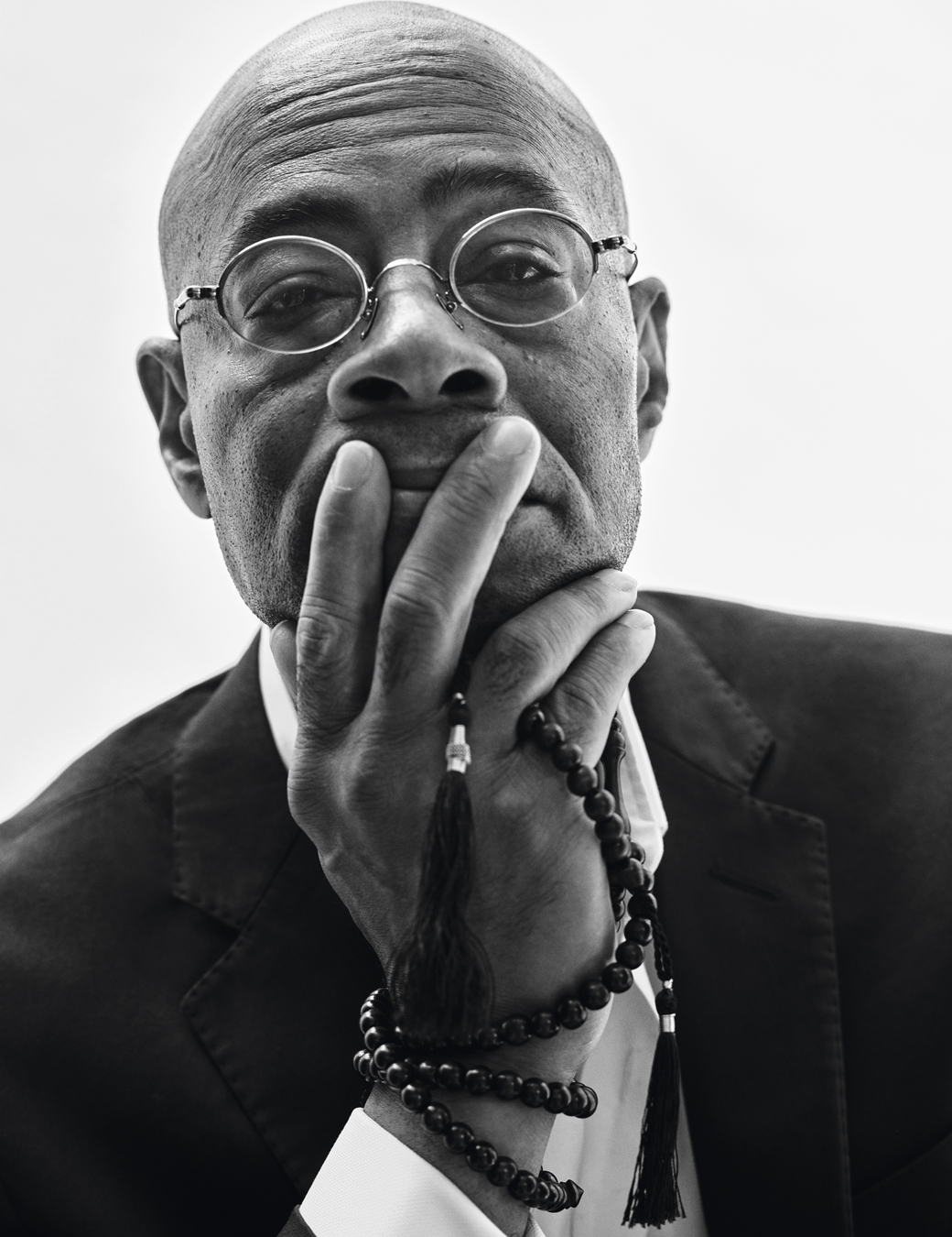 Model wears a white shirt, circular glasses and black blazer with rosary beads wrapped around his hand.