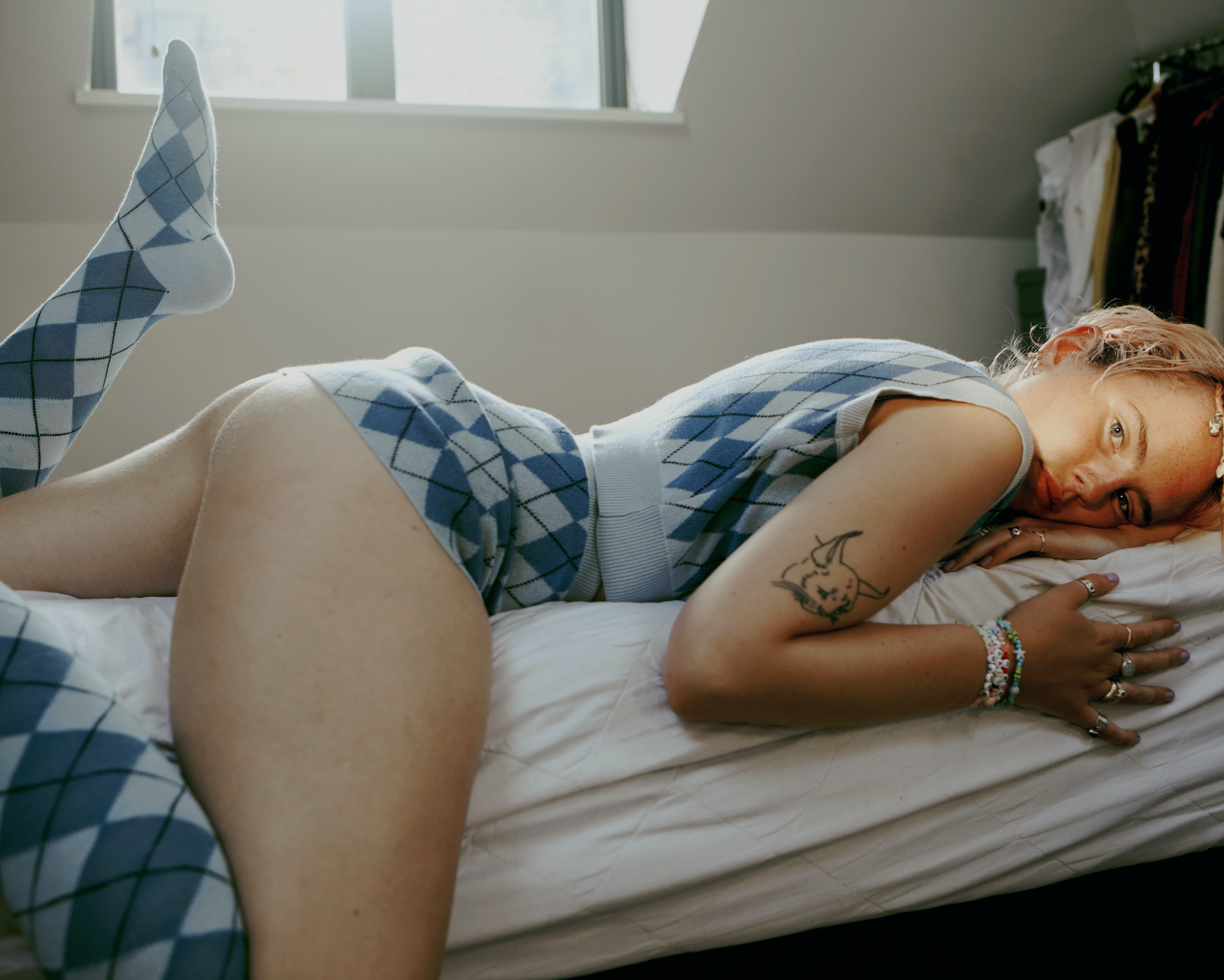 a girl in a knitted skirt and vest two-piece lays front down on her bed, sunlight on her face looking to camera