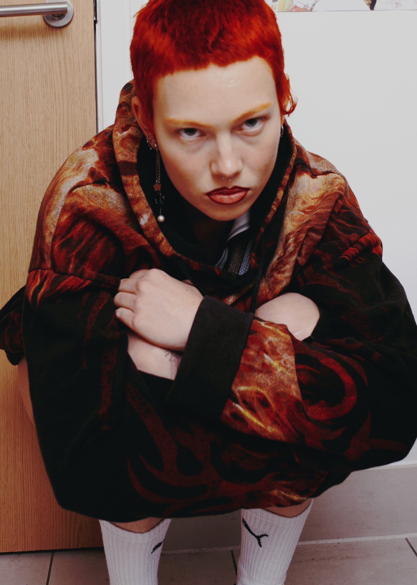 a girl with super short bright red hair and lined lips squats on a tiled floor