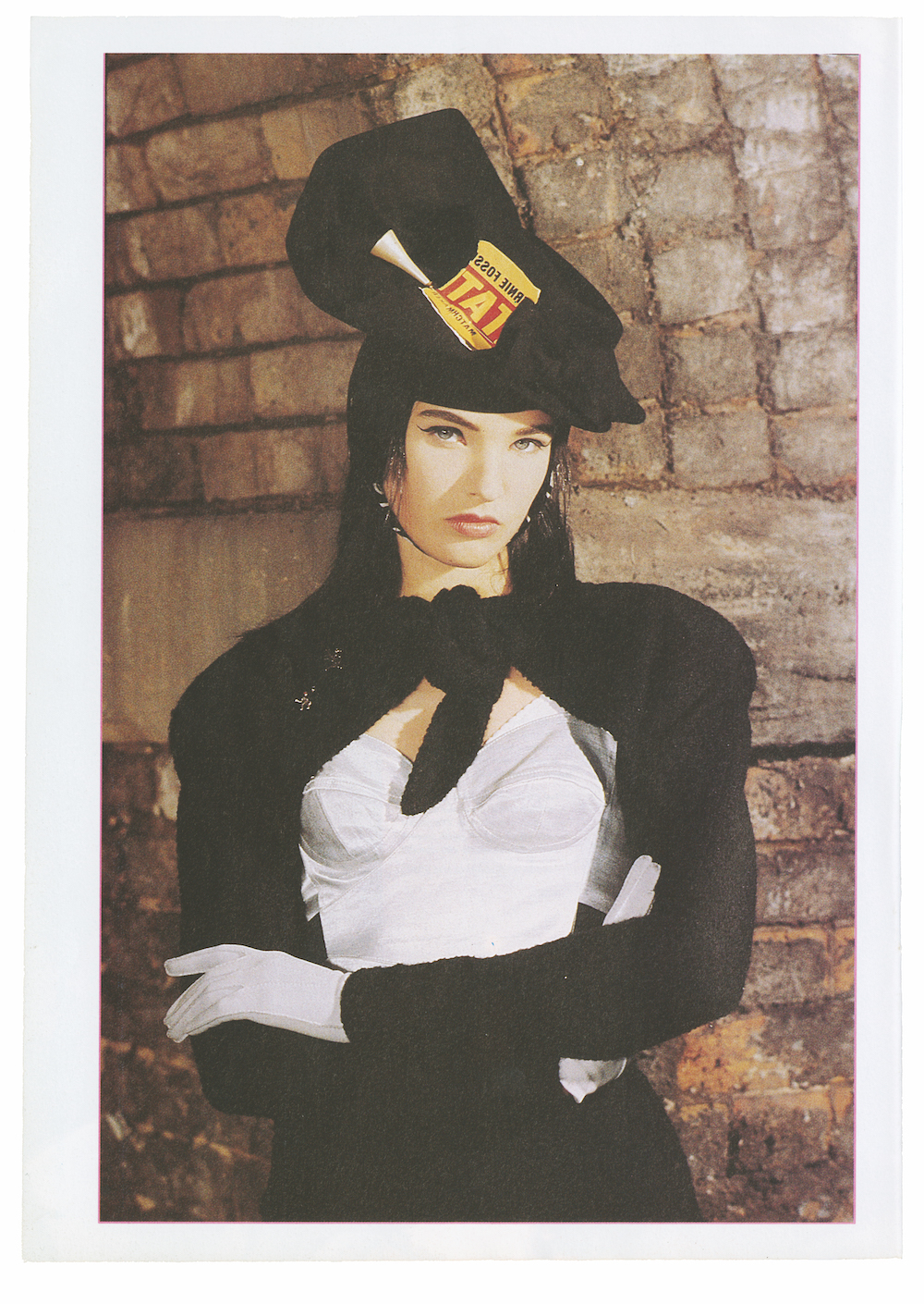 a woman in a black blouse, white corset and white gloves, wearing a hat with a torn-up piece of paper upon it