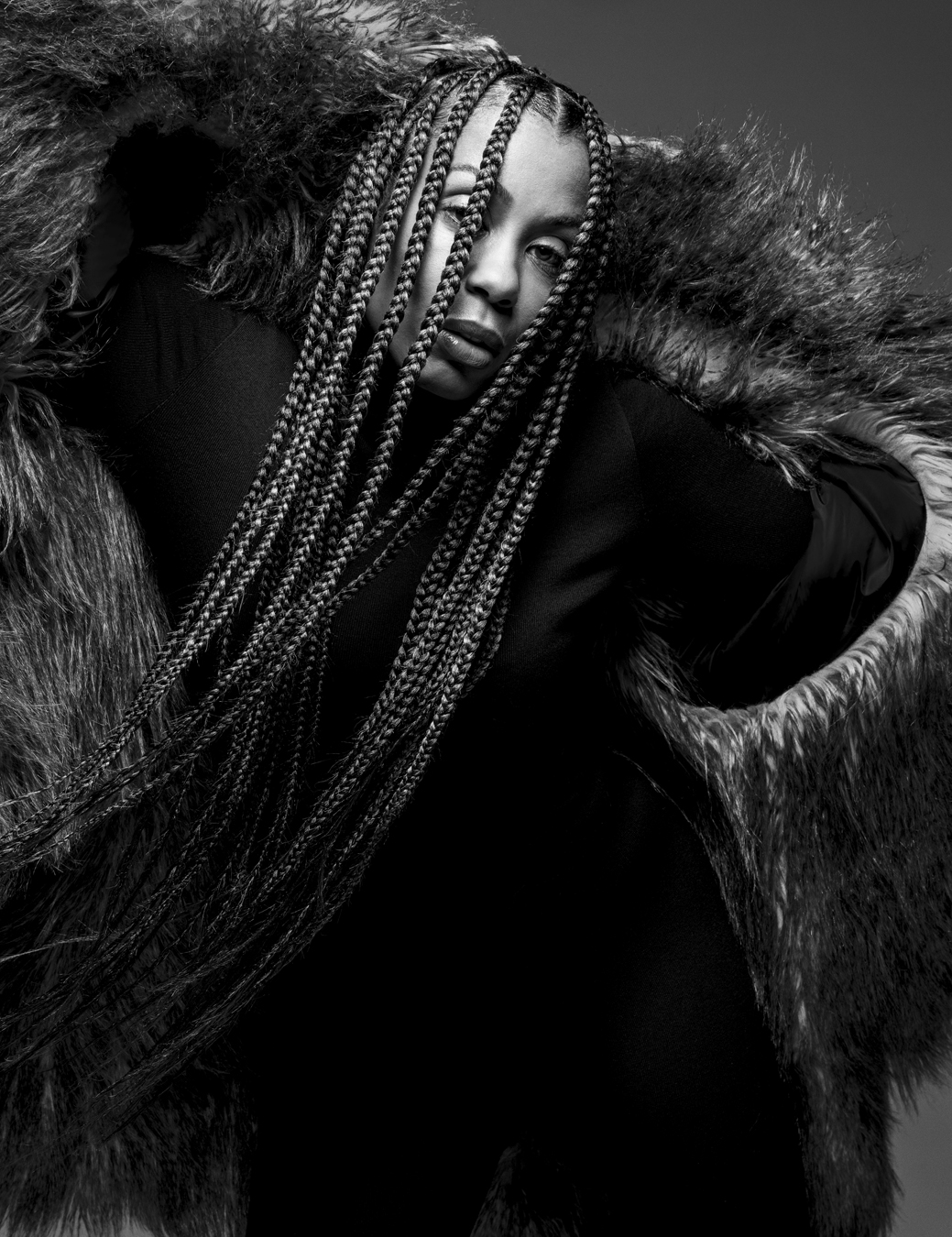 Portrait of woman wearing a black dress with a furry overcoat. Her braids fall across her face.