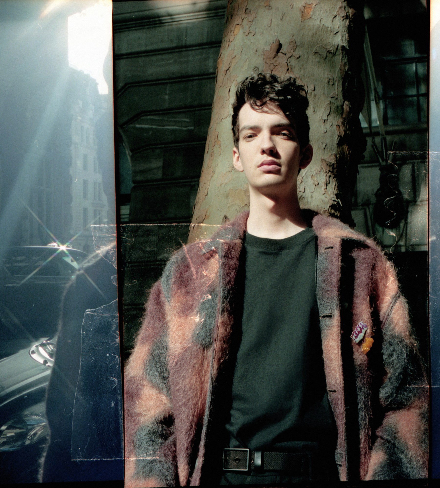 kodi smit mcphee in a mohair jacket and black t shirt leaning against a tree