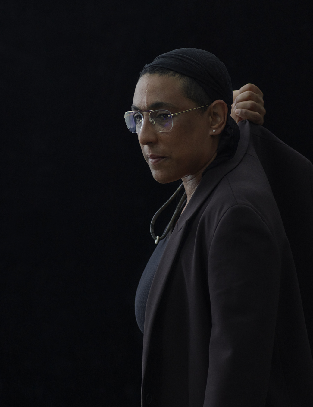 Leila stands against a black backdrop wearing a black top, a black blazer and a black do-rag.