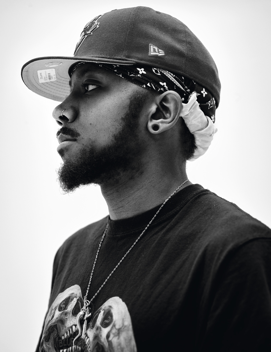 A side facing portrait of Mànsa. He wears a t-shirt with a two skulls print, a chain crucifix and snake necklace, a head tie and a baseball hat.