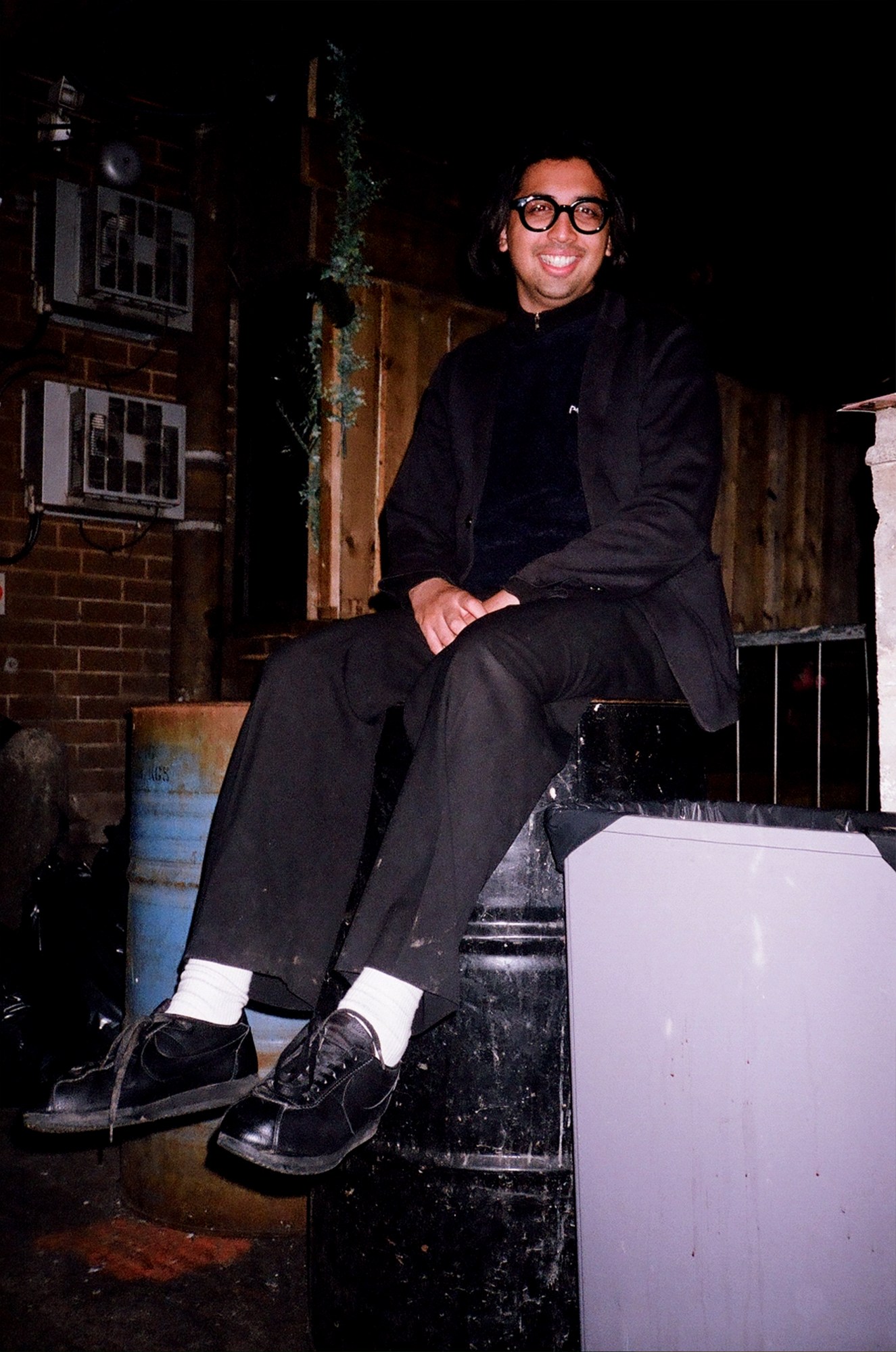 a guy with thick round glasses sits on top of a barrel in a smoking area, smiling to camera