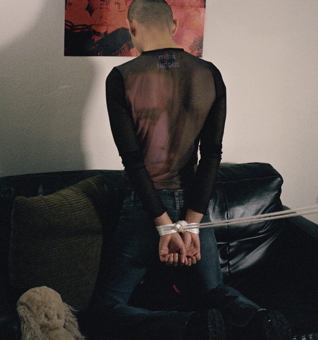 a young male-presenting person with their wrists tied behind their back kneels on a leather sofa, facing away from the camera