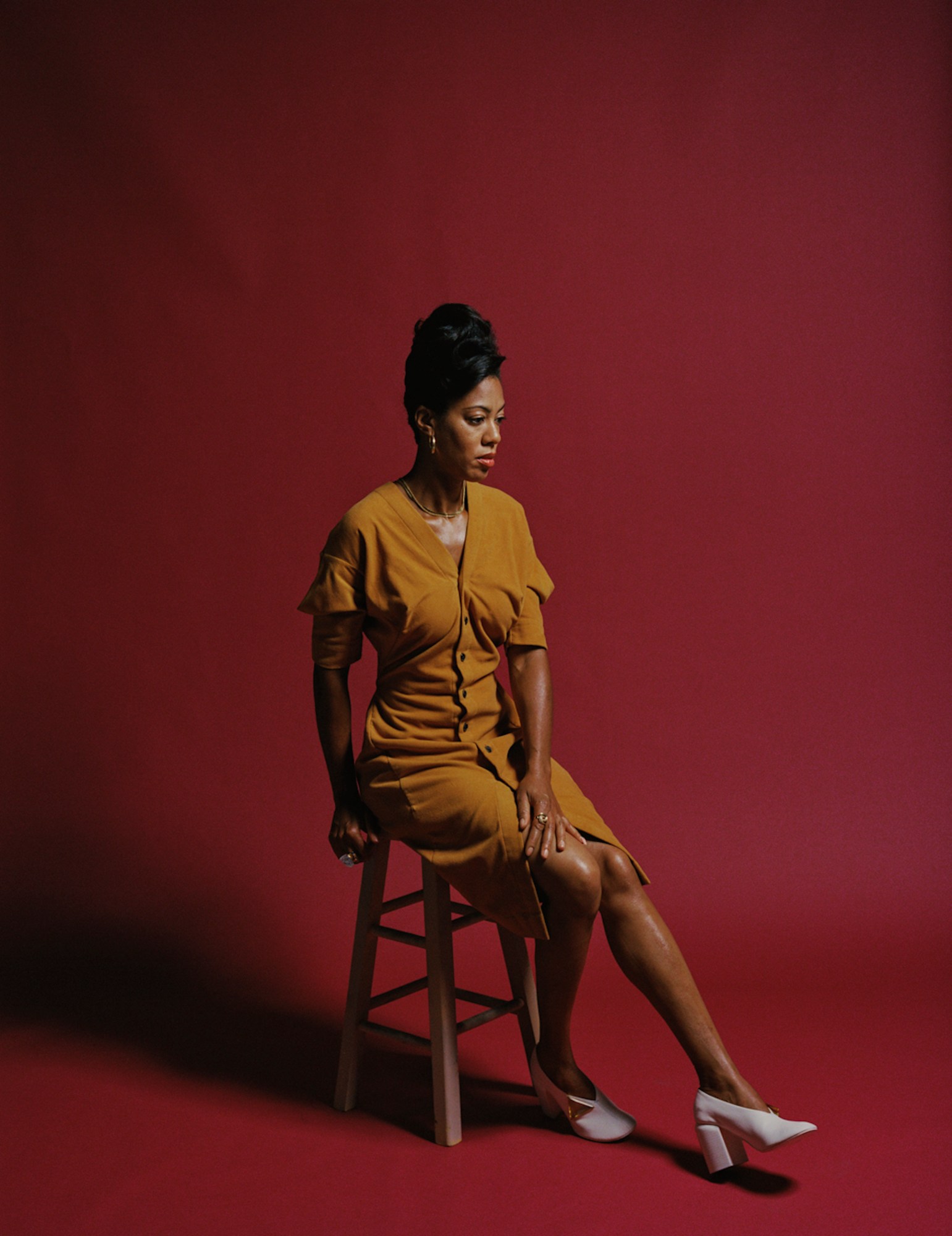 Deana sits on a stool wearing a yellow dress, white high heels, gold necklace, gold earrings and an up-do hairstyle.