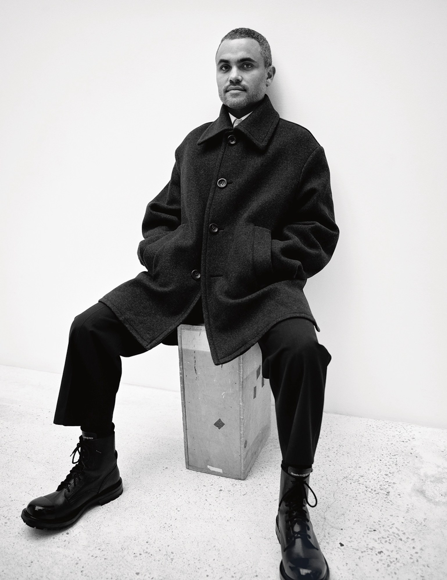 Thomas wears a black coat, trousers and boots as he sits on a tall box in a white cemented room