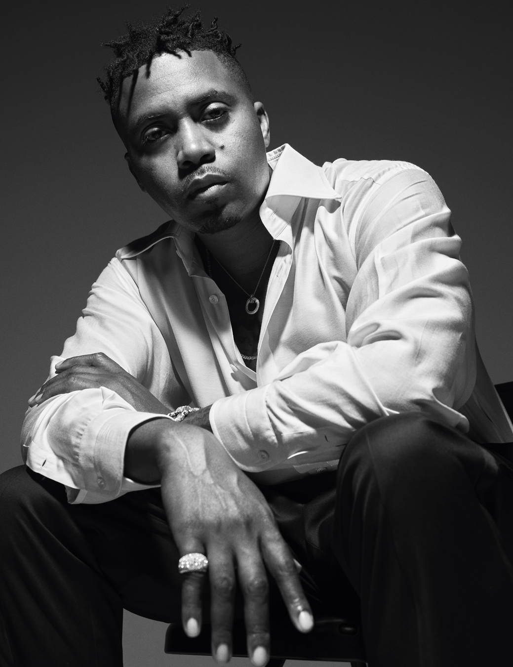 Nas sits wearing a white shirt and black trousers, a silver ring pendant chain, a chain bracelet and a silver ring.