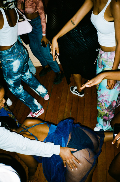 A group dancing with one person in the middle in a sheer blue dress twerking while someone grabs her butt.