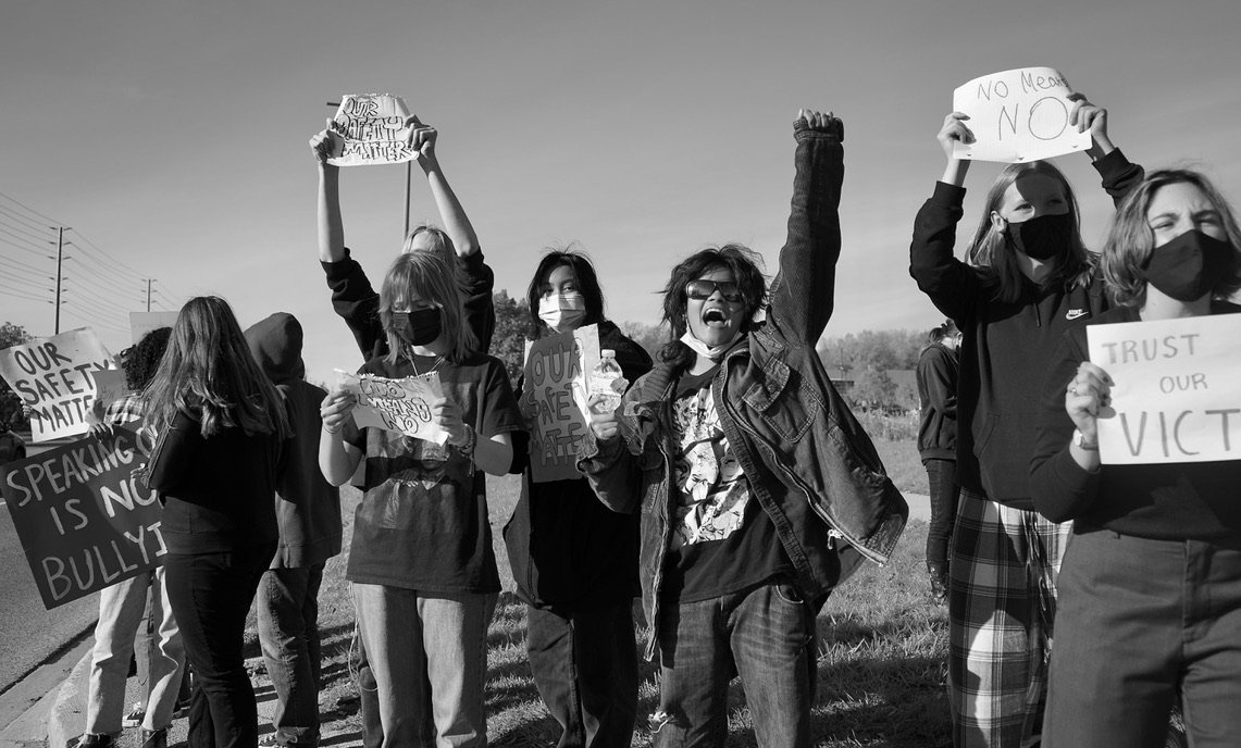 A group of female protesters wearing masks and holding signs stand by the edge of a road.