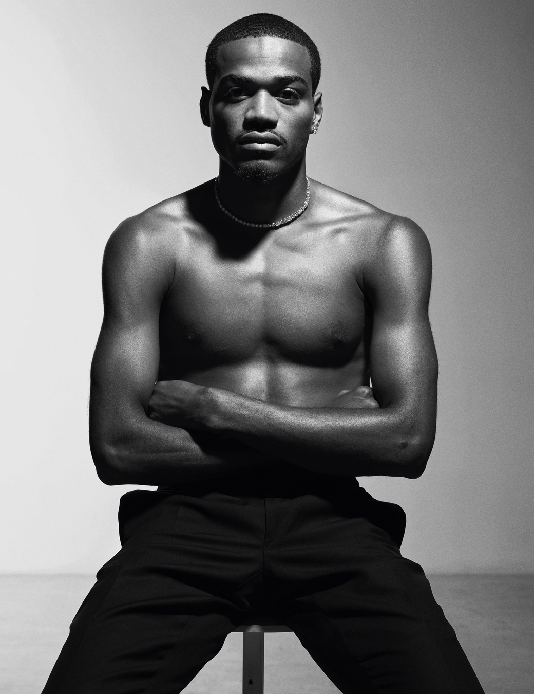 Tyshawn sitting on a stool shirtless wearing a diamond necklace and black tailored trousers.