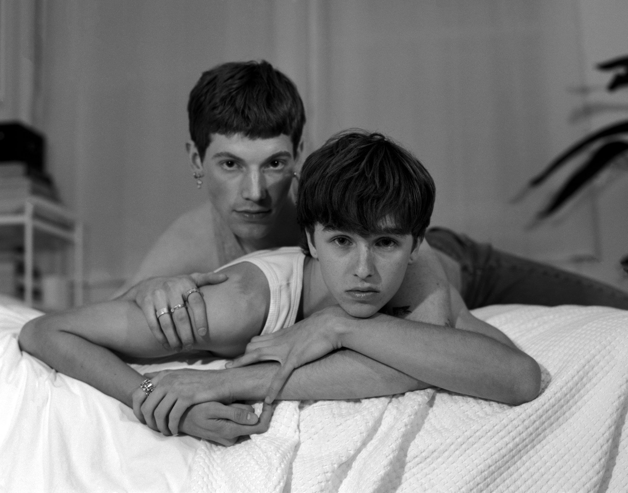 Two boys, one shirtless the other wearing a white vest lie on a bed.