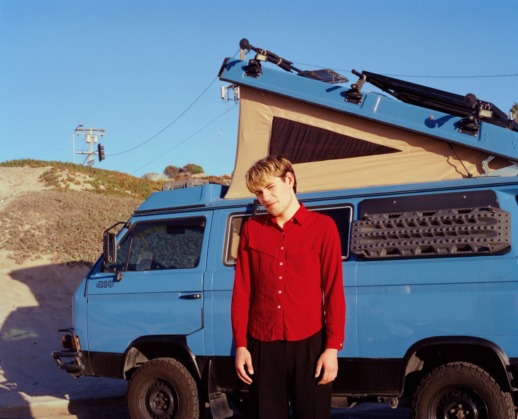 A boy in a red shirt stands in front of a camper van in the desert.