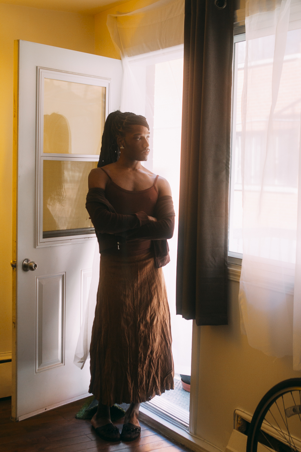 A girl stands in the doorway of a home wearing a brown dress and brown cardigan.