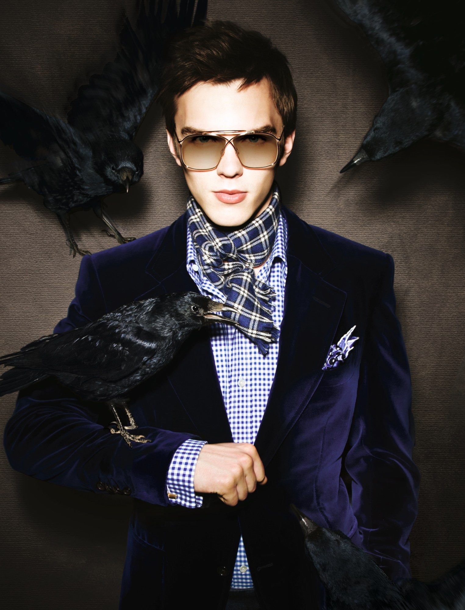 Nicholas Hoult wearing a blue check shirt, blue velvet blazer a check neck scarf and dark sunglasses with a raven on his arm.