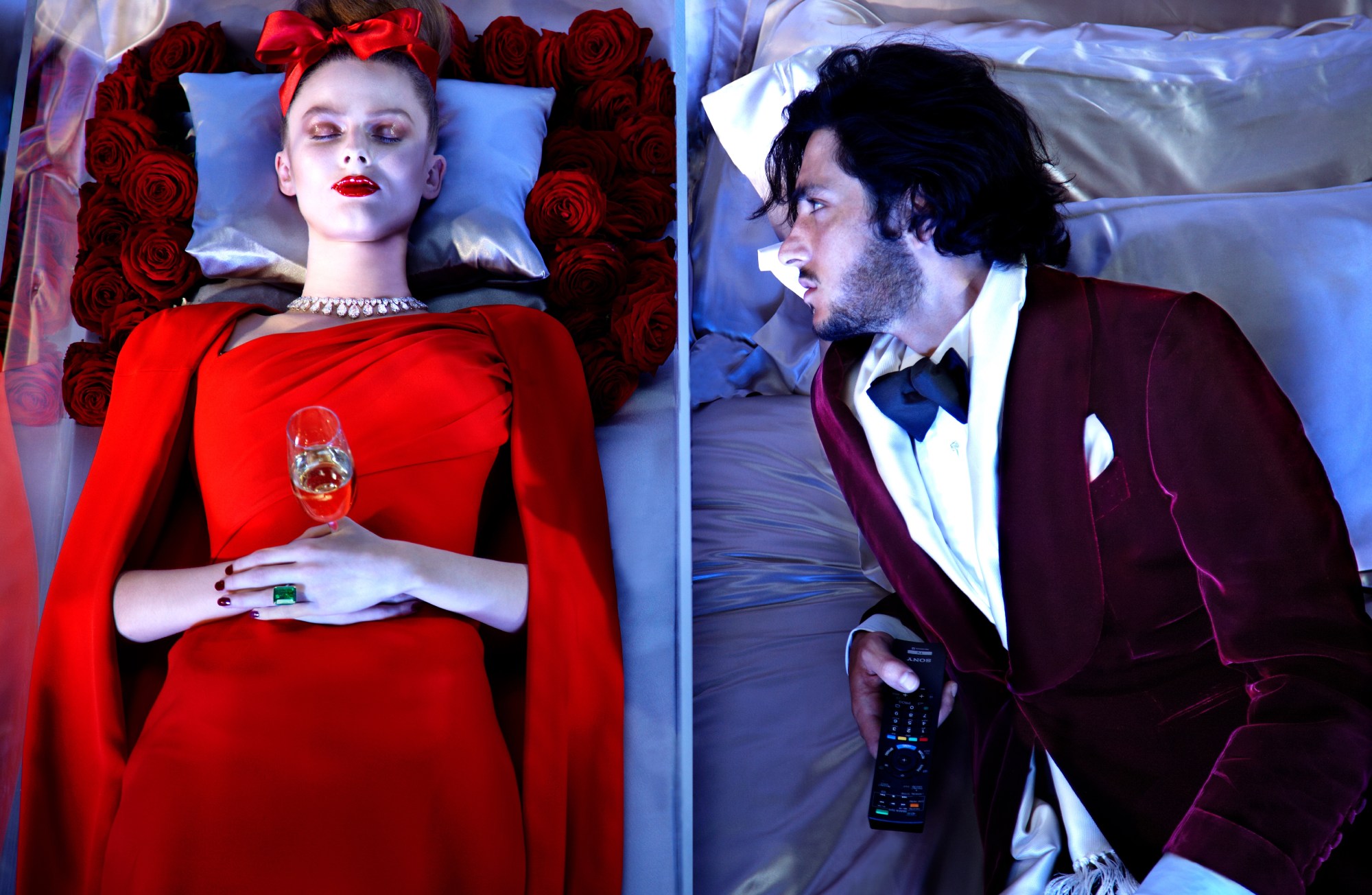 Woman wearing red and holding a champagne glass lies in a coffin. Next to her is a man in a burgundy velvet suit staring at her and holding a remote.