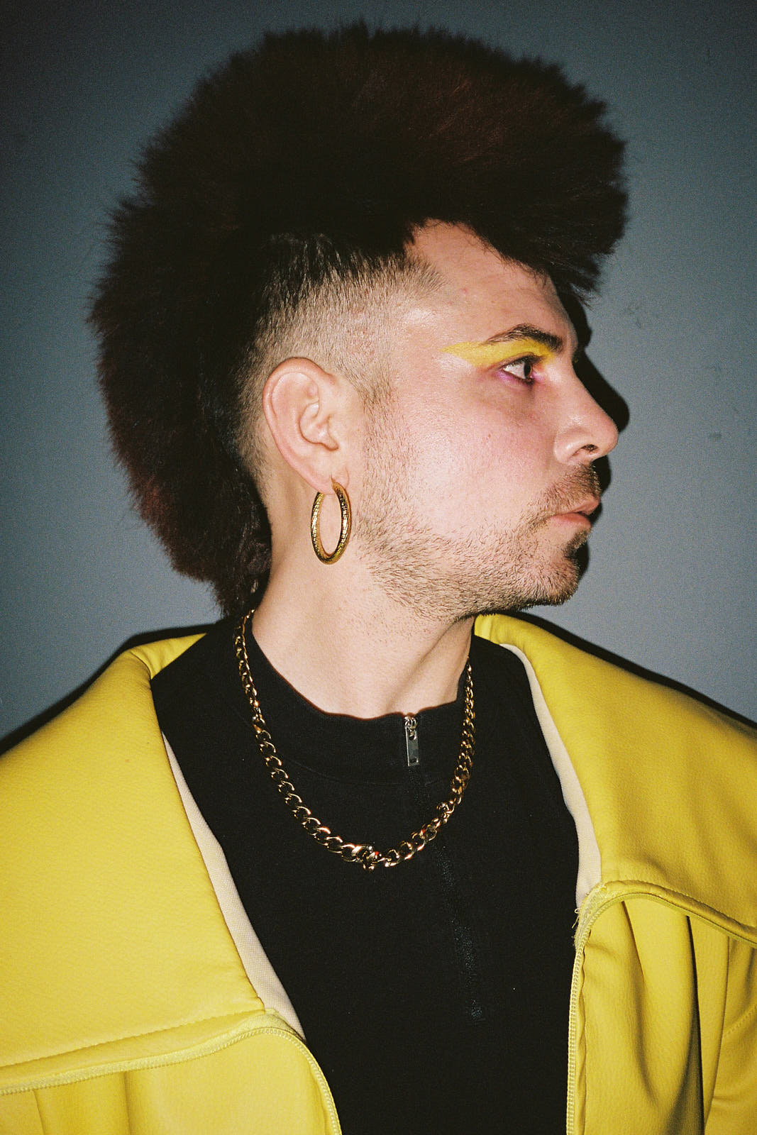 a close up of a person with a fluffy mohawk wearing a bright yellow jacket and yellow eyeshadow