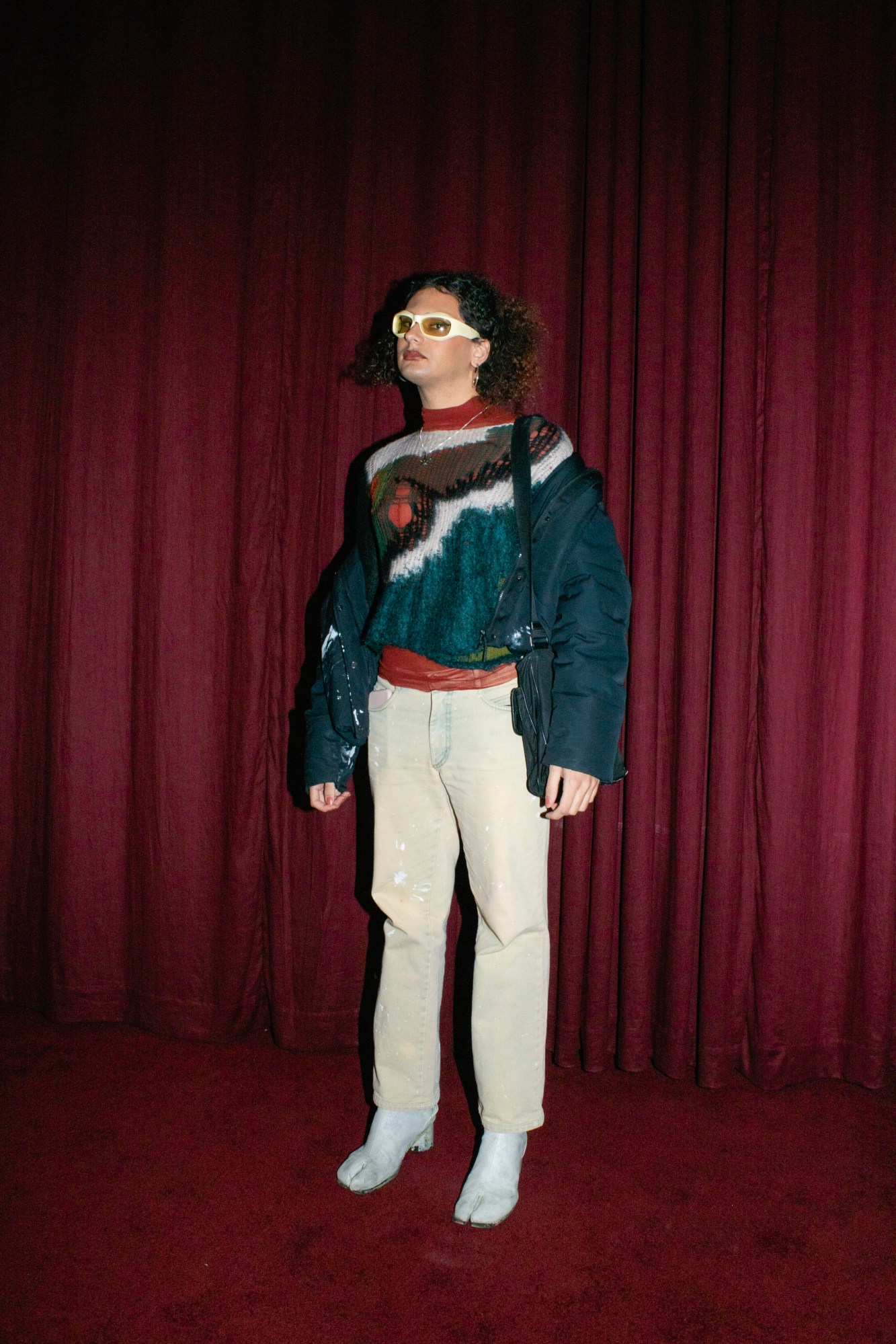 a person with a colourful knit jumper, pale jeans and tabi boots stands in front of a red curtain