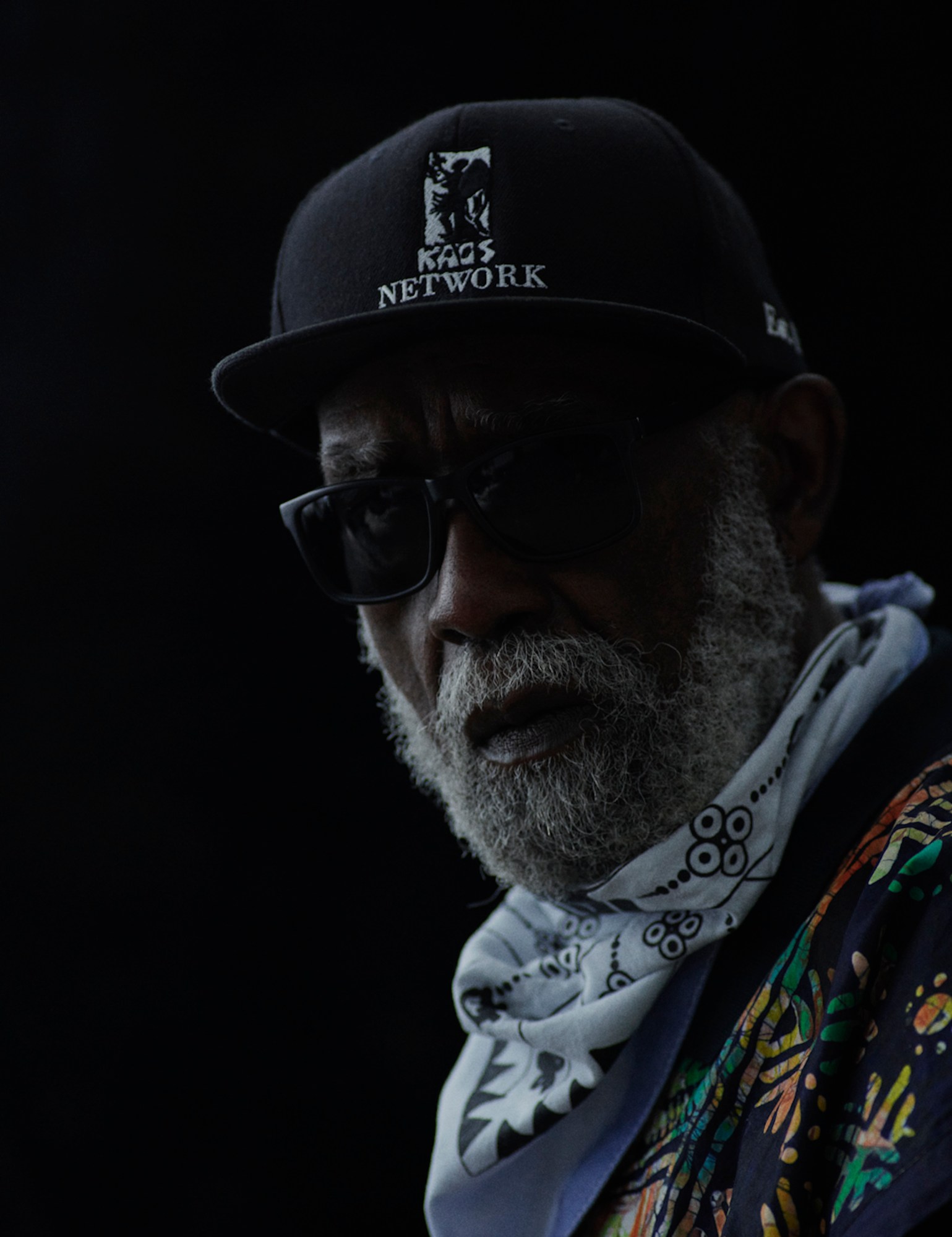 Portrait of ben wearing a colourful floral print shirt with a light blue scarf, blackout sunglasses and a black cap.