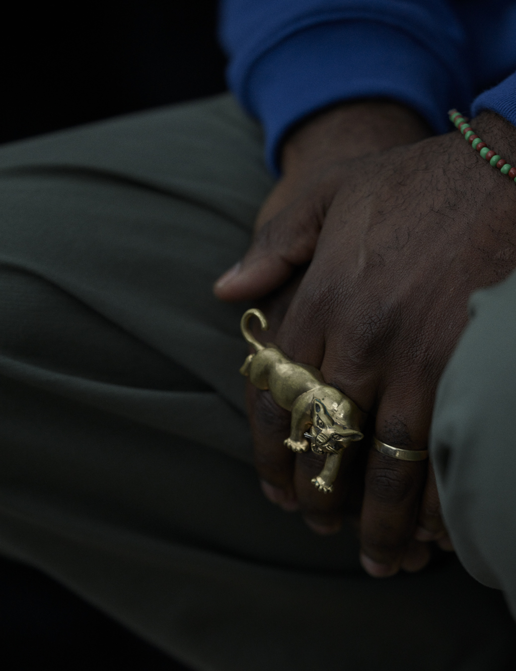 A close up of Kahlil's hands on his lap with a lion ring across two fingers and a gold ring on another and a beaded bracelet on his wrist.