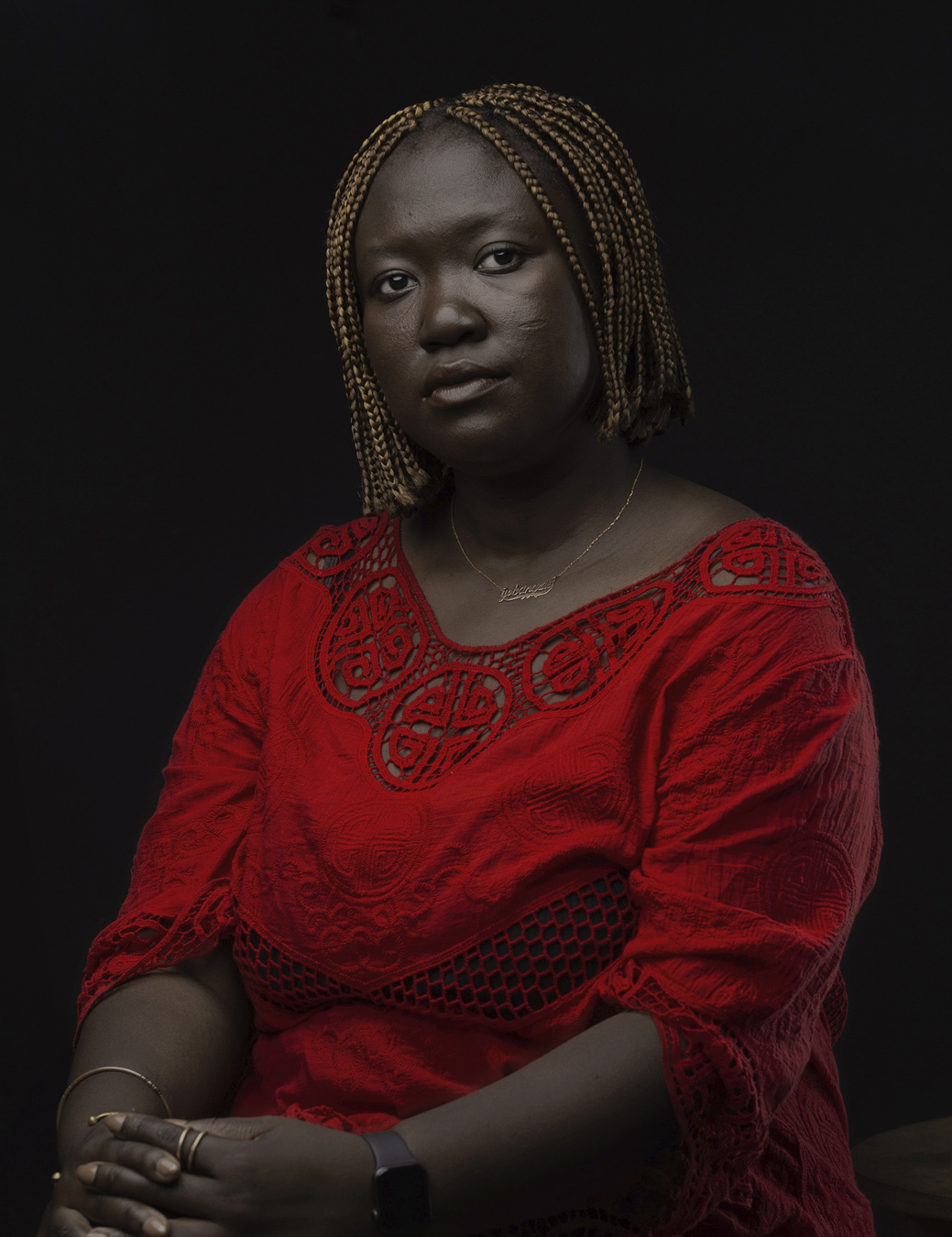 Nuotama sits wearing a red lacy cut out dress with a gold word necklace and gold bracelets and rings.