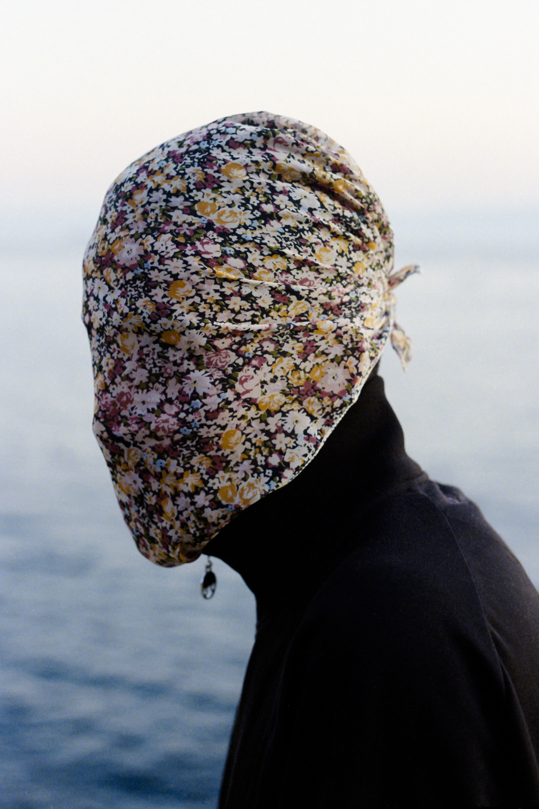 a portrait of a man with a floral scarf over his face in front of the ocean