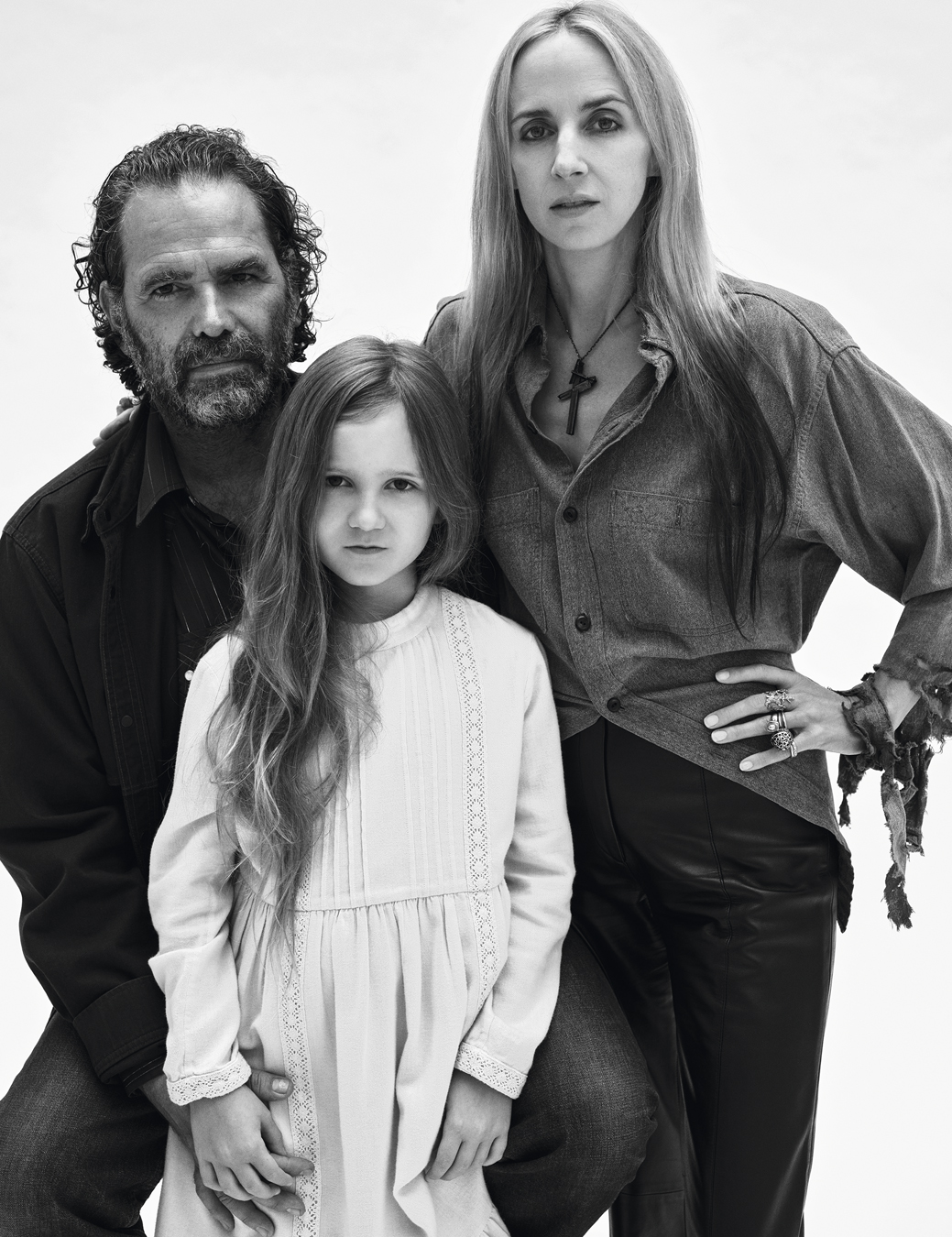 Gavin wearing all black and Hope wearing a grey blouse, a crucifix necklace and strips of fabric on her arm, stand with their daughter in a white dress.