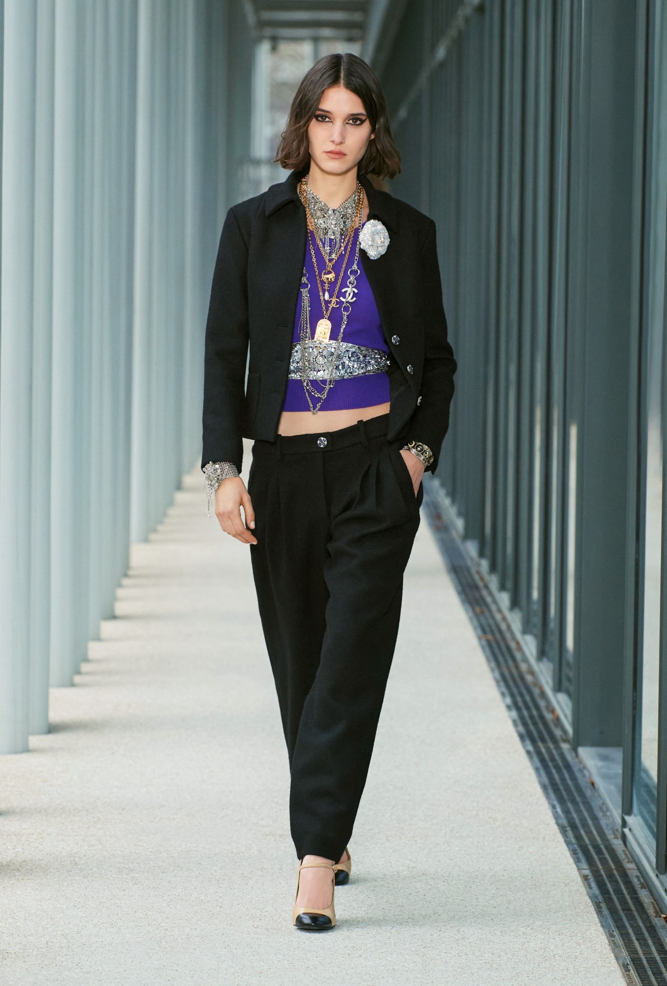 Model wears black tailored trousers and jacket with a purple cropped top, a silver belt and lots of necklaces with a flower broach.