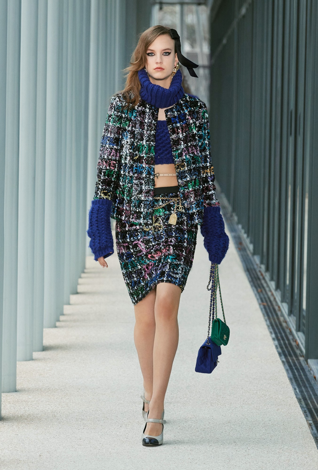 Model wears a tweed two piece suit with royal blue arm warmers, snood and bandeau top and holds two mini handbags.